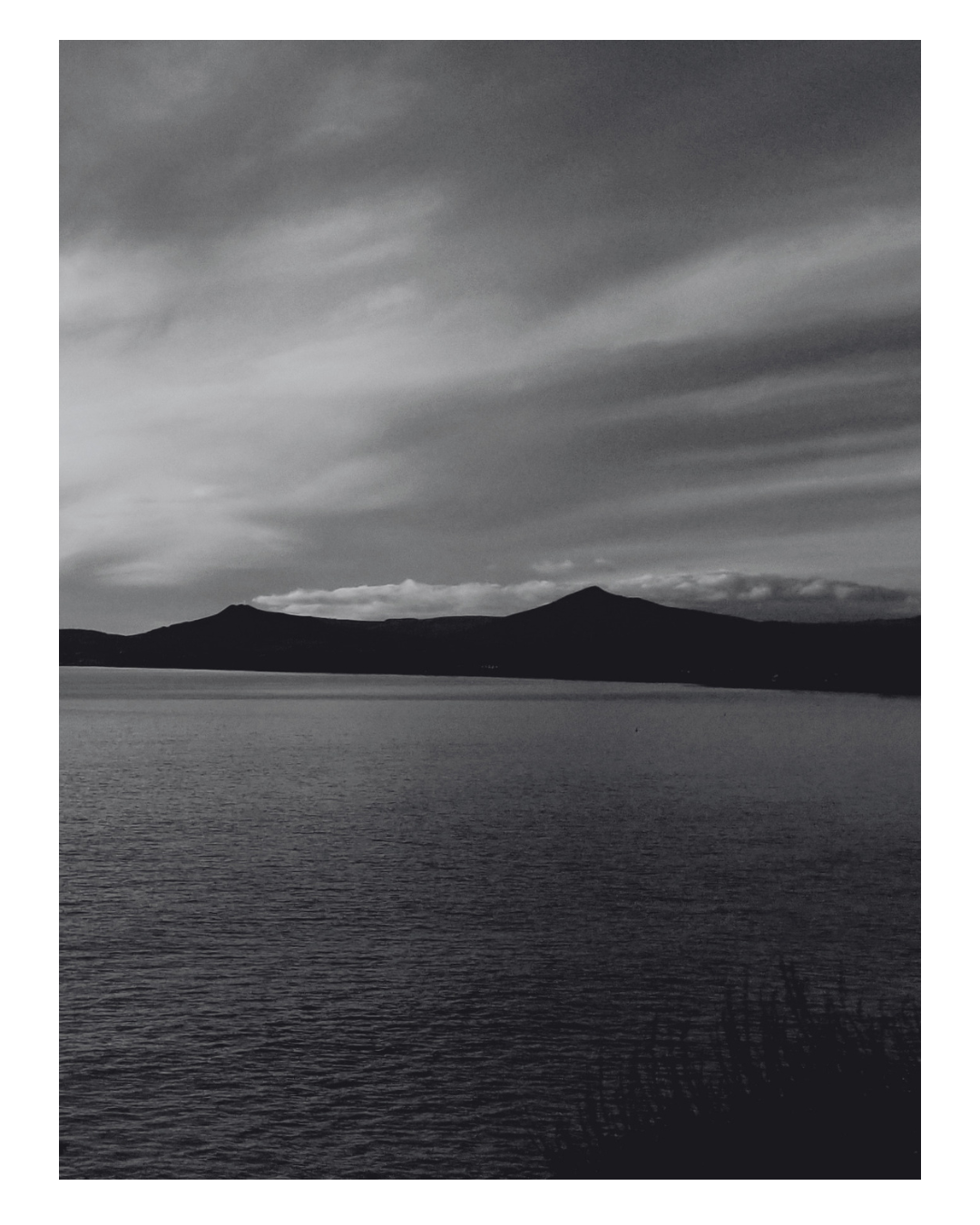 Black and white photograph of a calm lake with mountain range in the background and a cloudy sky above.