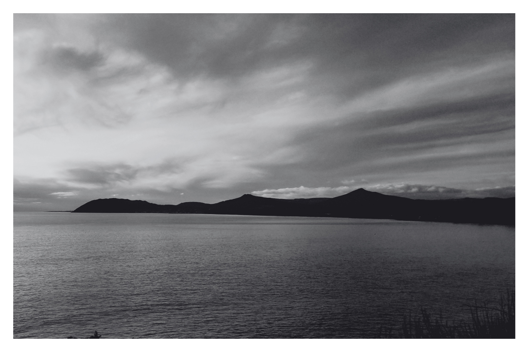 Black and white photo of a large body of water with distant hills and mountains under a cloudy sky.