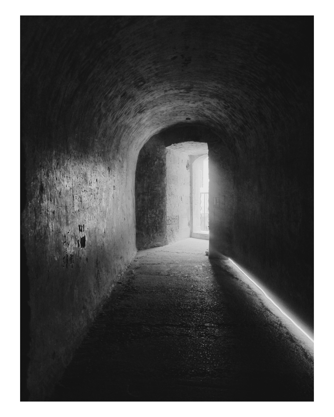 Dark arched tunnel leading to a brightly lit doorway with a window, black and white photograph.