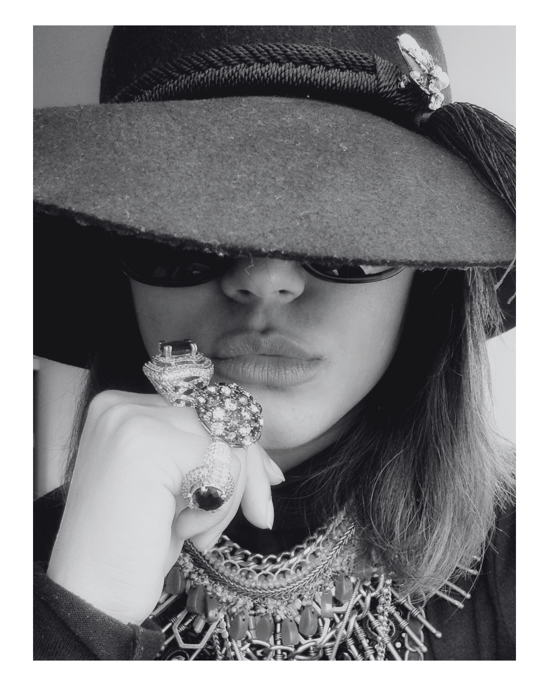 Woman wearing a large brimmed hat with a butterfly brooch, dark sunglasses, and multiple layered necklaces, holding a large ornate ring near her lips in black and white.