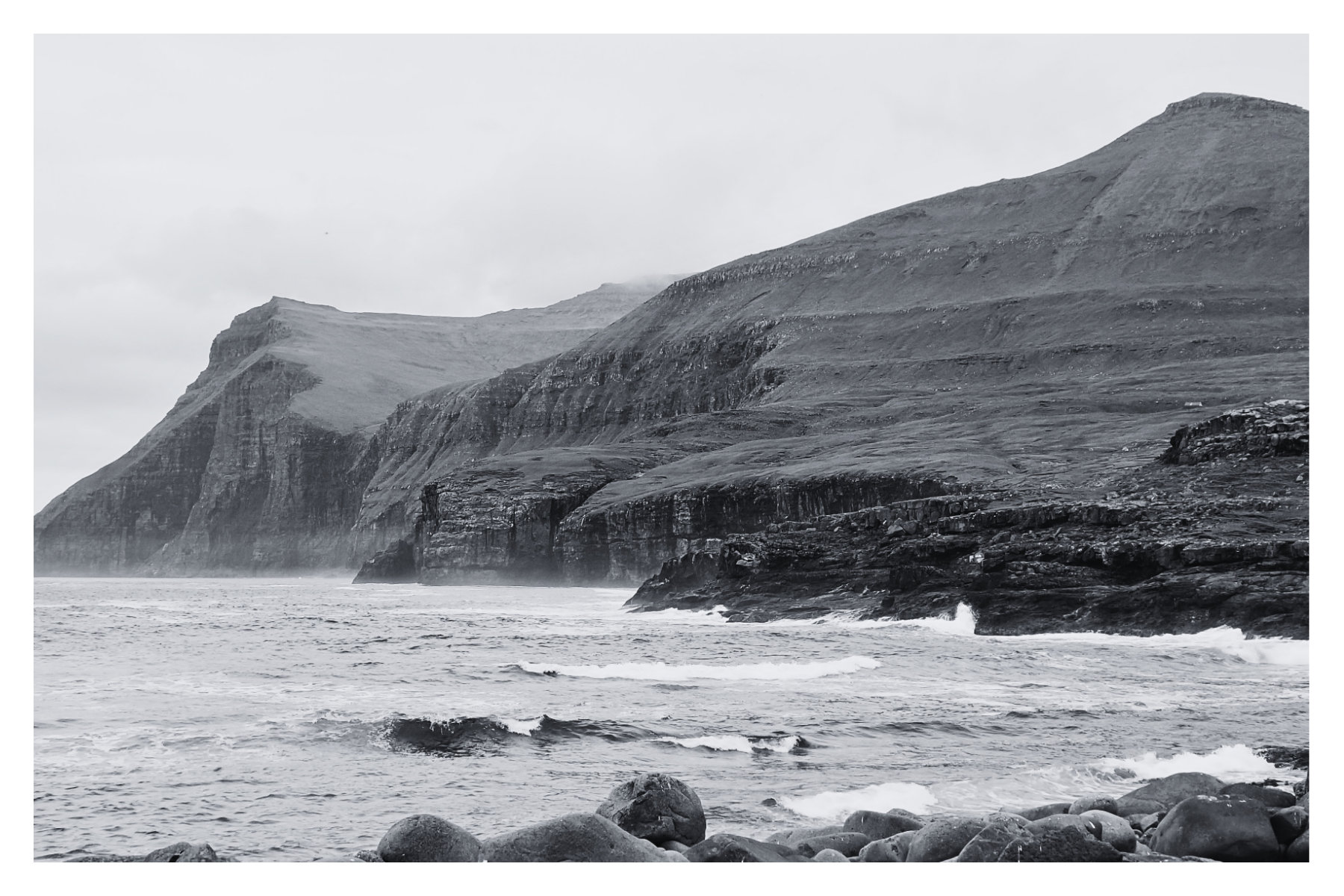 Black and white photo of a rugged coastline with steep cliffs and rough ocean waves crashing against rocks.