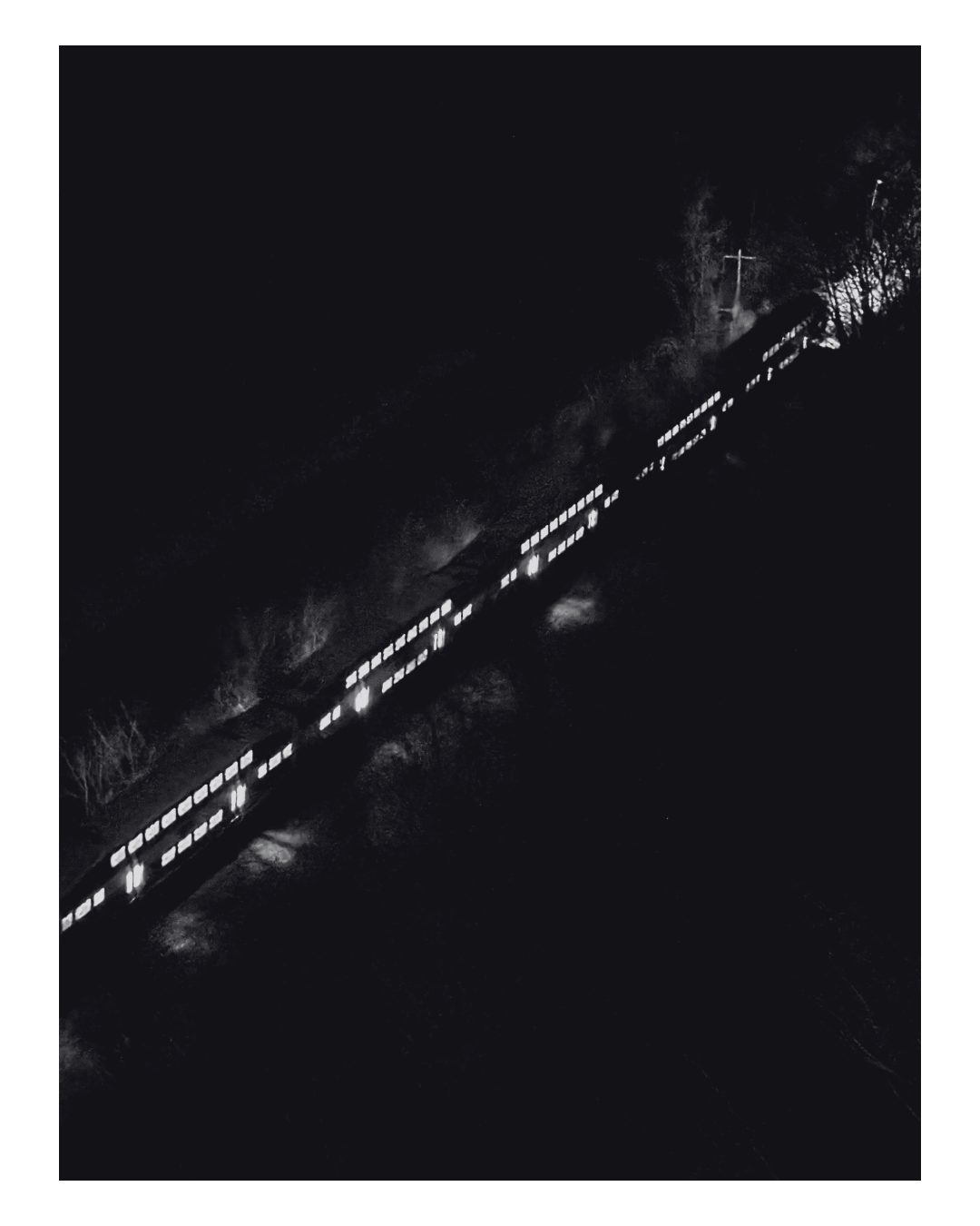 Aerial night view of a train with illuminated windows traveling on tracks through a dark landscape with some trees and a cross on a hill in the background.