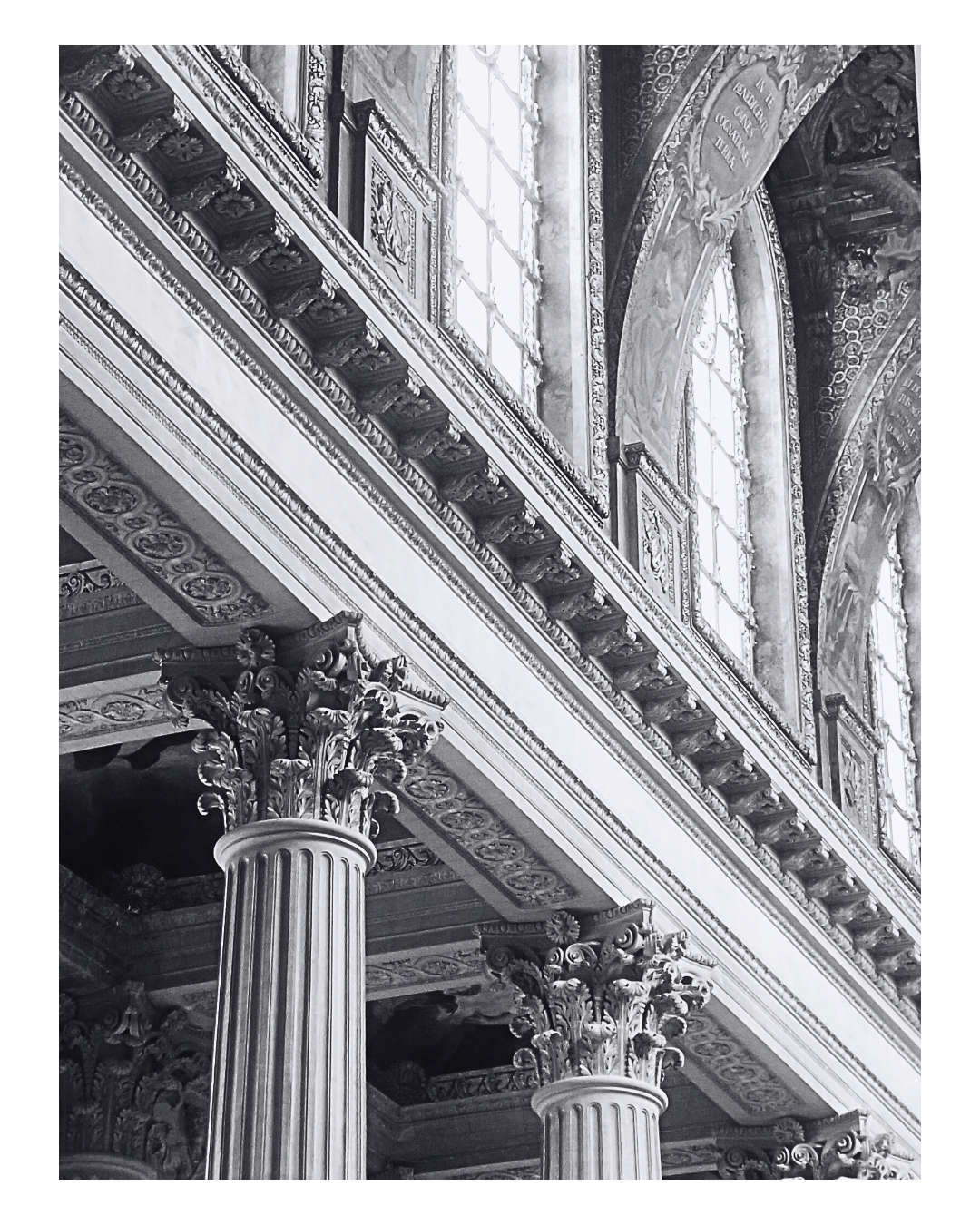 Black and white photograph of ornate architectural details, including large classical columns with intricate capitals, decorative friezes, and elaborate window frames with multiple rectangular panes.