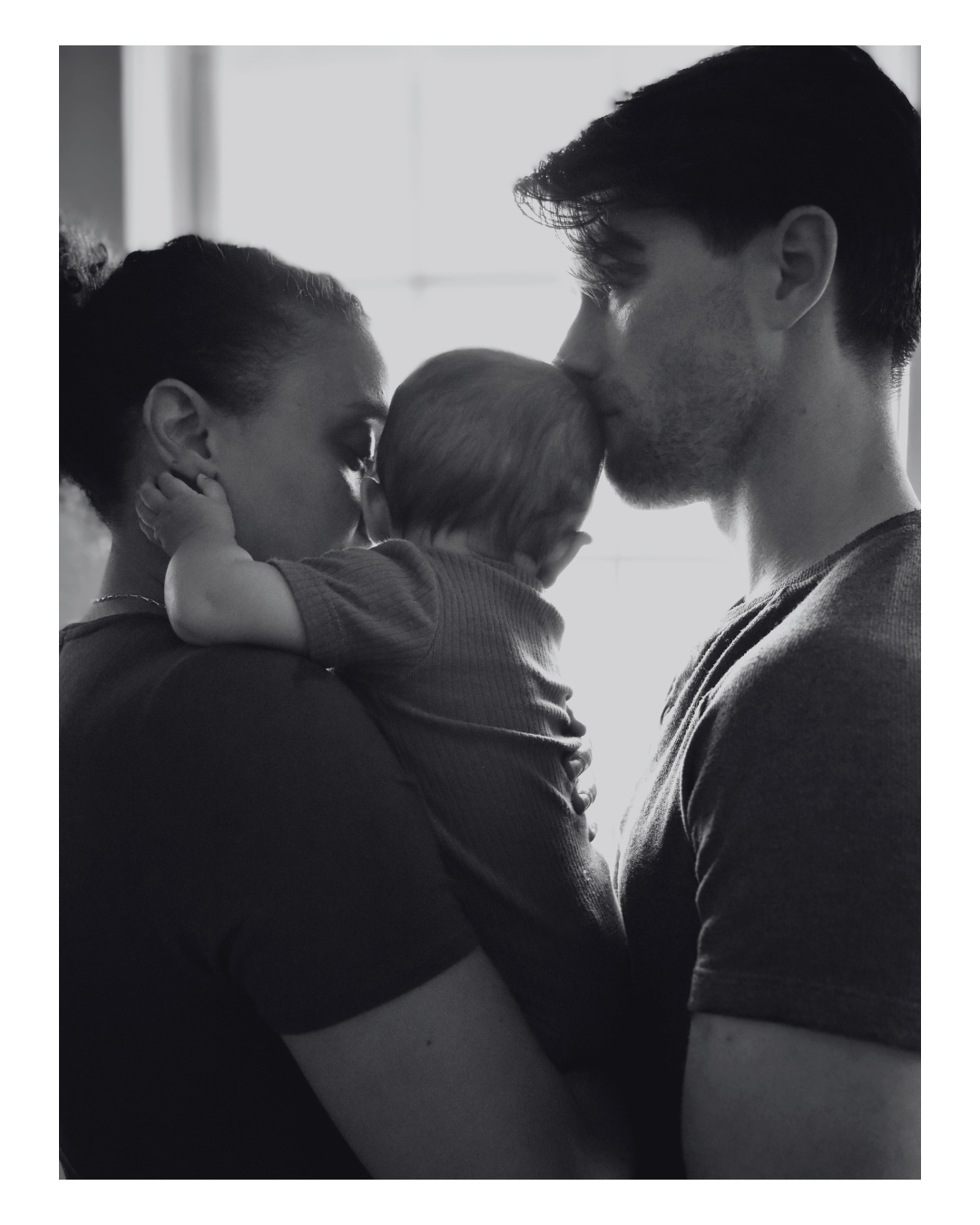 Black and white photo of a family with two adults and a baby, the adults kissing the baby's head, backlit by a window.