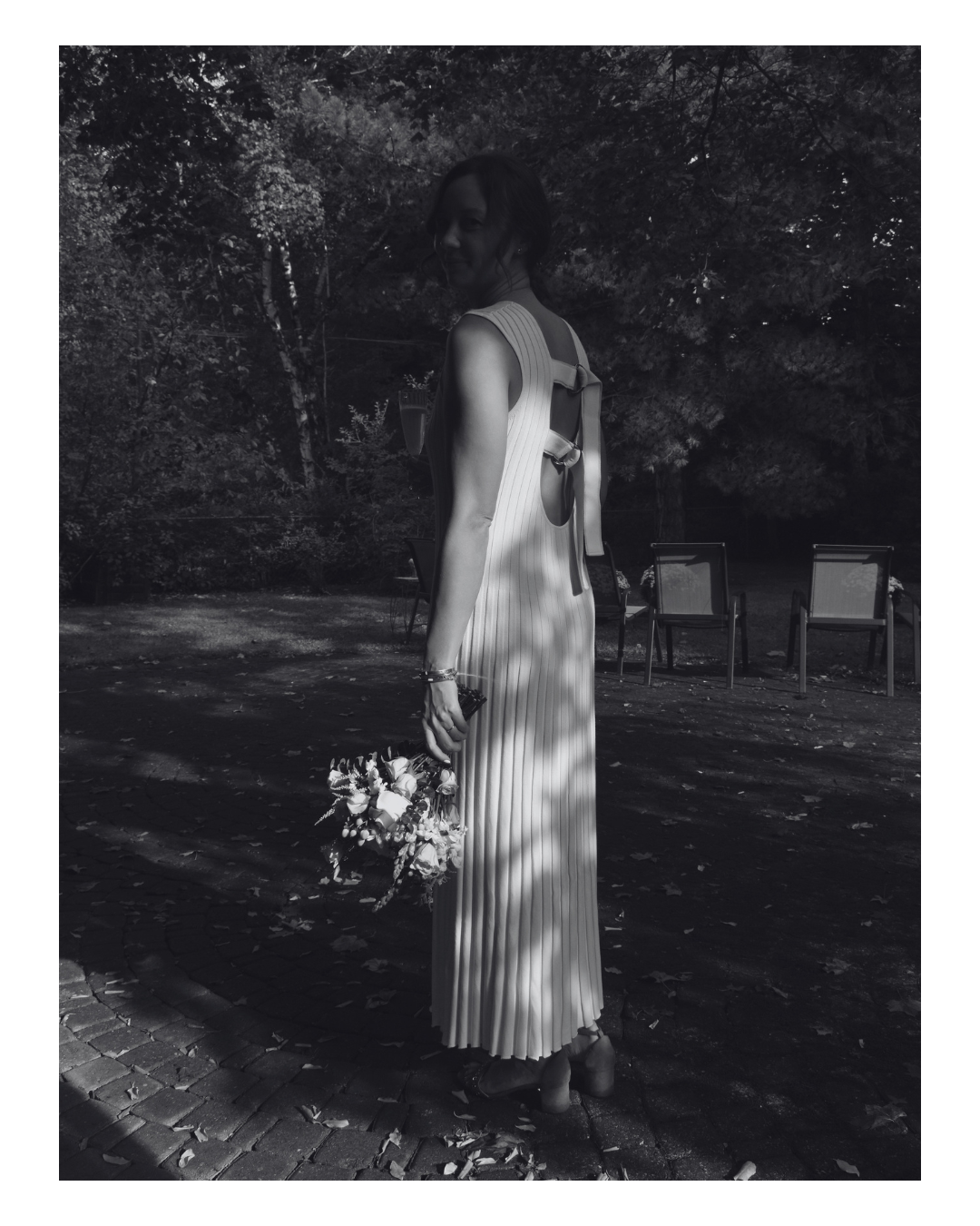 A woman standing outdoors at night, wearing a sleeveless dress and holding a bouquet of flowers, with trees and outdoor chairs in the background.