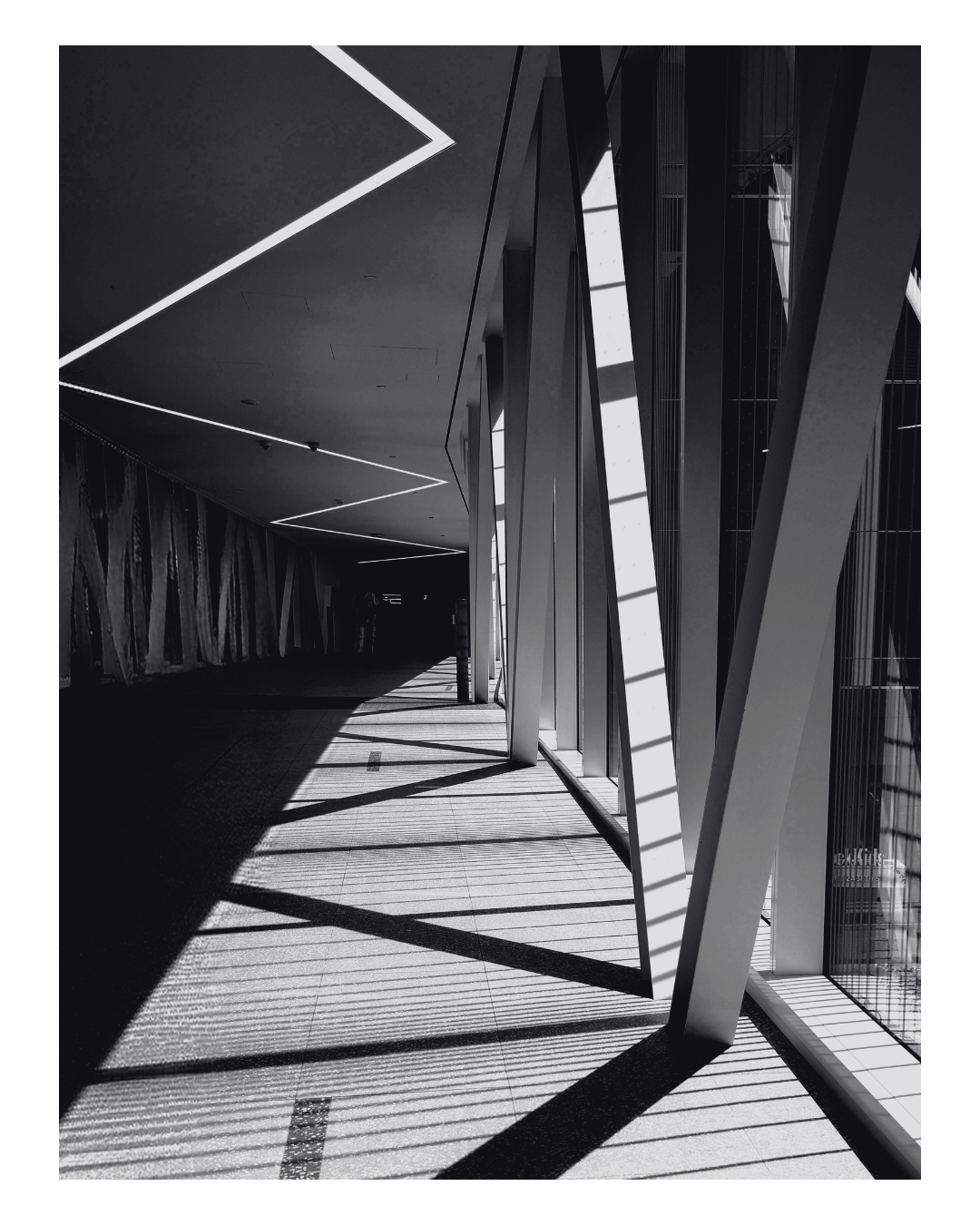 Black and white photo of a modern building corridor with large glass windows and angular architectural elements. Sunlight casts shadow patterns on the floor.