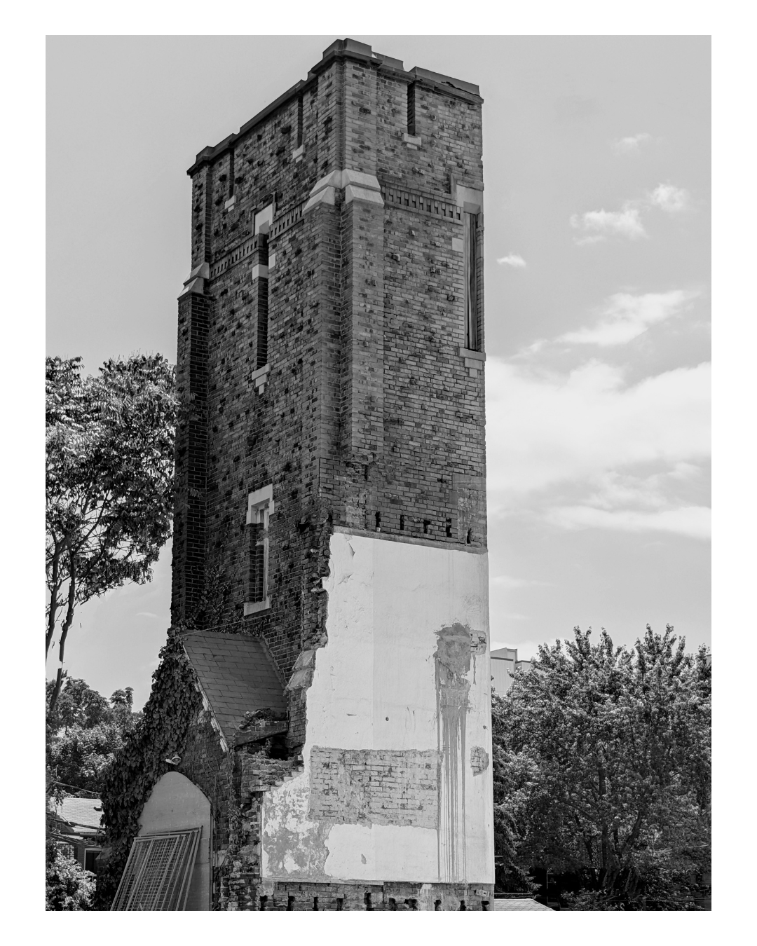 Black and white photo of a tall, partially damaged brick building or tower with windows, surrounded by trees.