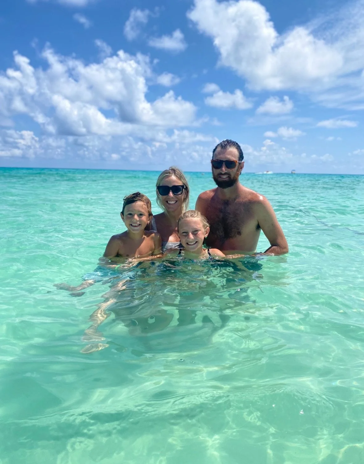 A family of four, including a man, woman, and two children, smiling and enjoying swimming in clear turquoise water at the beach under a partly cloudy sky.