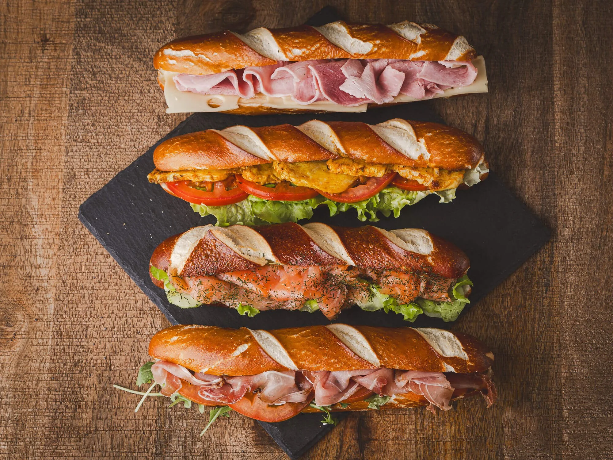 Quatre sandwiches de baguette avec différentes garnitures, tous présentés sur une plaque noire sur une surface en bois.