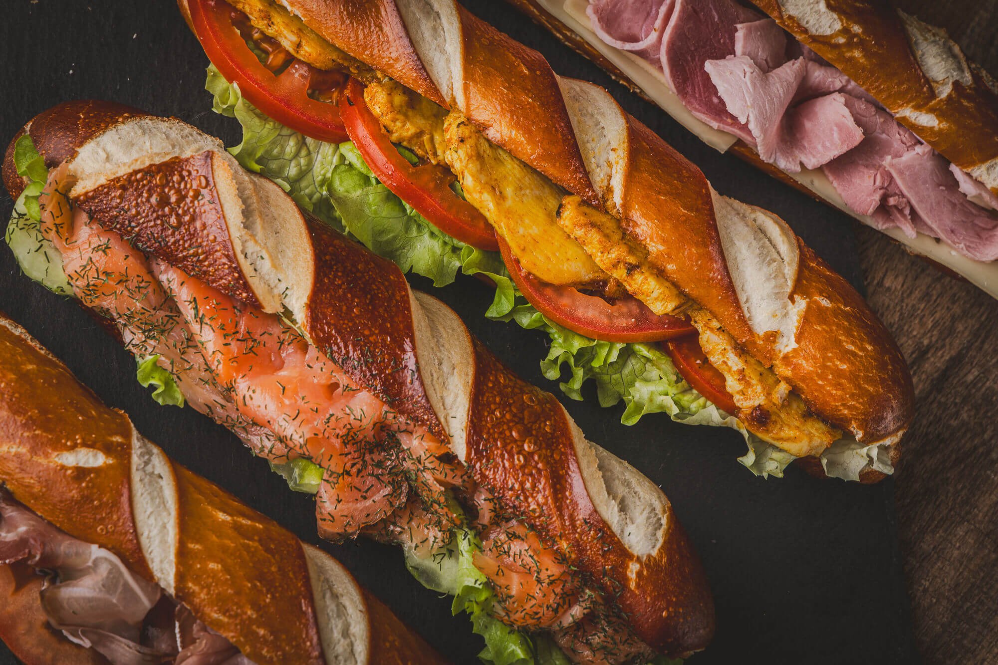 Trois sandwichs fusettes variés avec des ingrédients frais, légumes et viandes, servis sur une planche en bois.