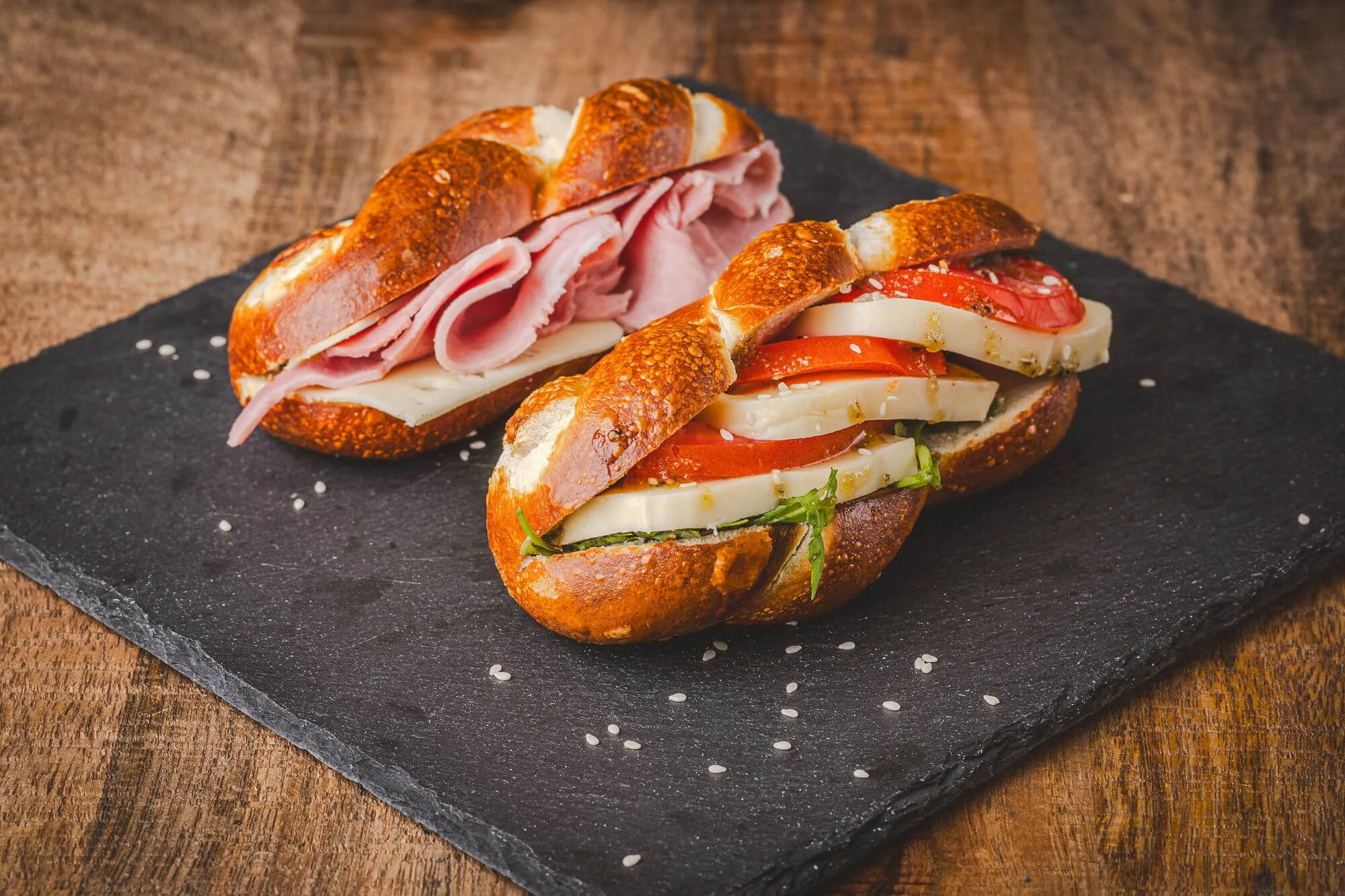 Deux sandwichs tresses bretzels sur une plaque de ardoise, garnis de jambon, fromage, tomates, laitue, avec des graines de sésame, sur une table en bois.
