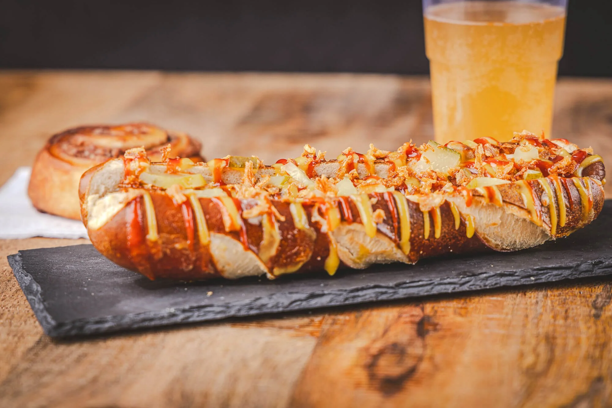Un hot dog de bretzel garni de ketchup, moutarde, oignons frits, et cornichons, avec un cinnamon roll et un verre de soda en arrière-plan.