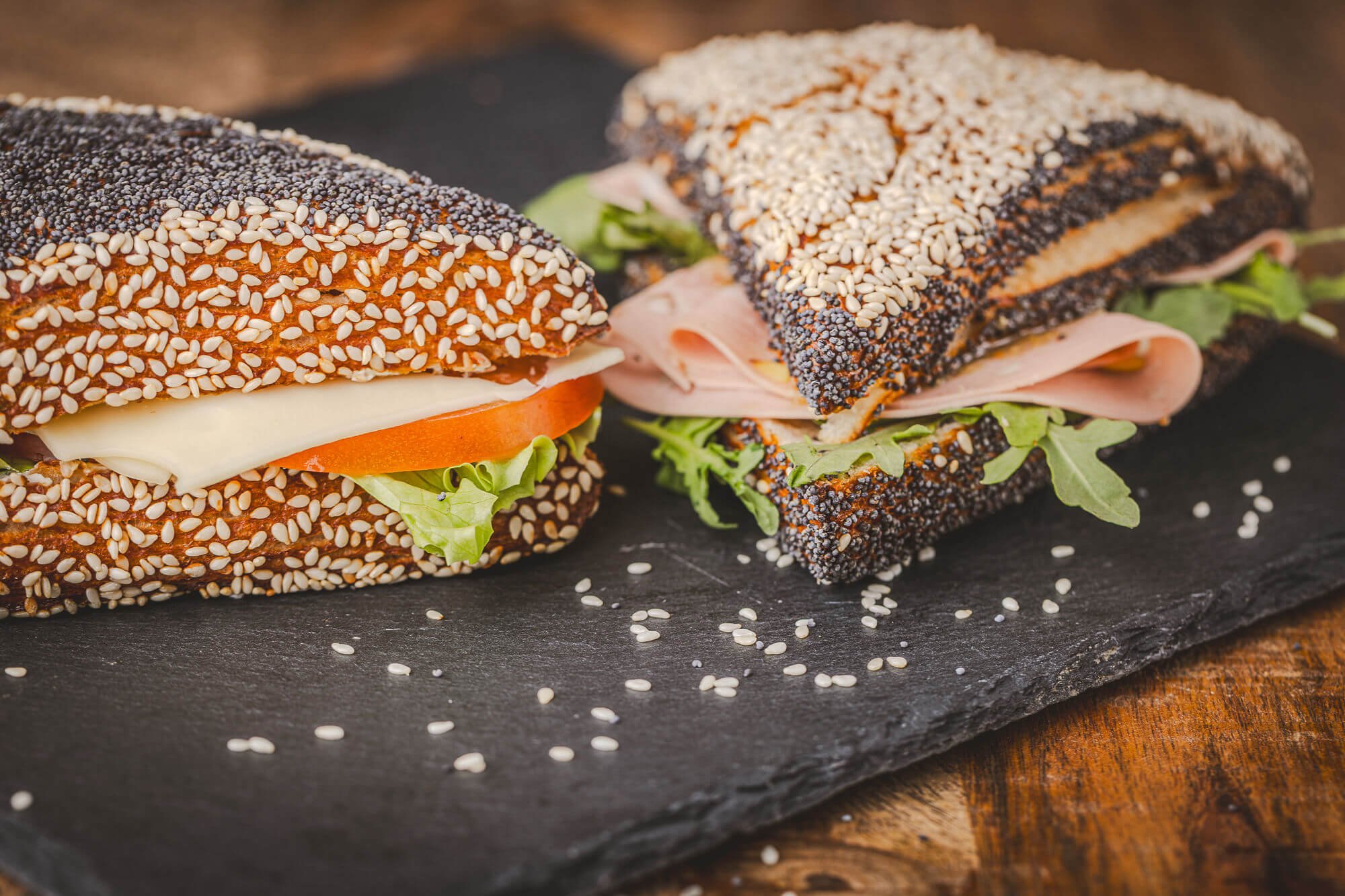 Sandwich triangle bretzel au pain aux graines de sésame avec jambon, fromage, tomate, laitue, présenté sur une planche en ardoise.