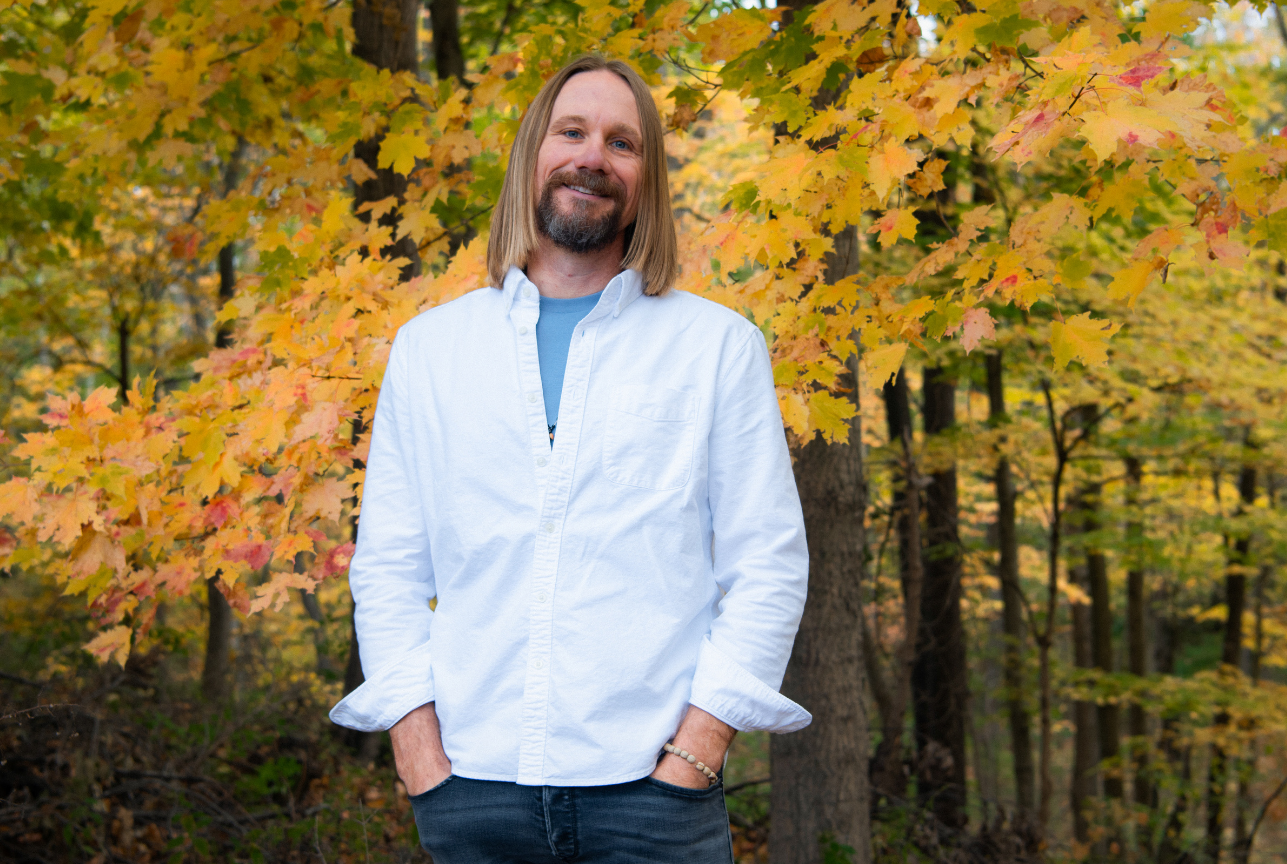 A man with shoulder-length blond hair and a beard, smiling, stands in front of autumn-colored trees in a forest, wearing a white shirt and blue t-shirt.