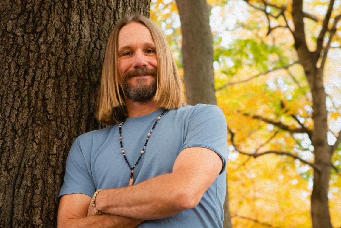 A man with shoulder-length blonde hair, a beard, and a mustache smiling as he leans against a tree in a forest with autumn-colored leaves.