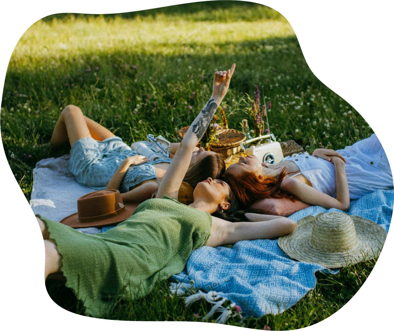 Three women lying on a blanket in a grassy field, relaxing and enjoying a picnic on a sunny day and doing playful, unique mindfulness activities for self-discovery, self-expression and deeper connection.