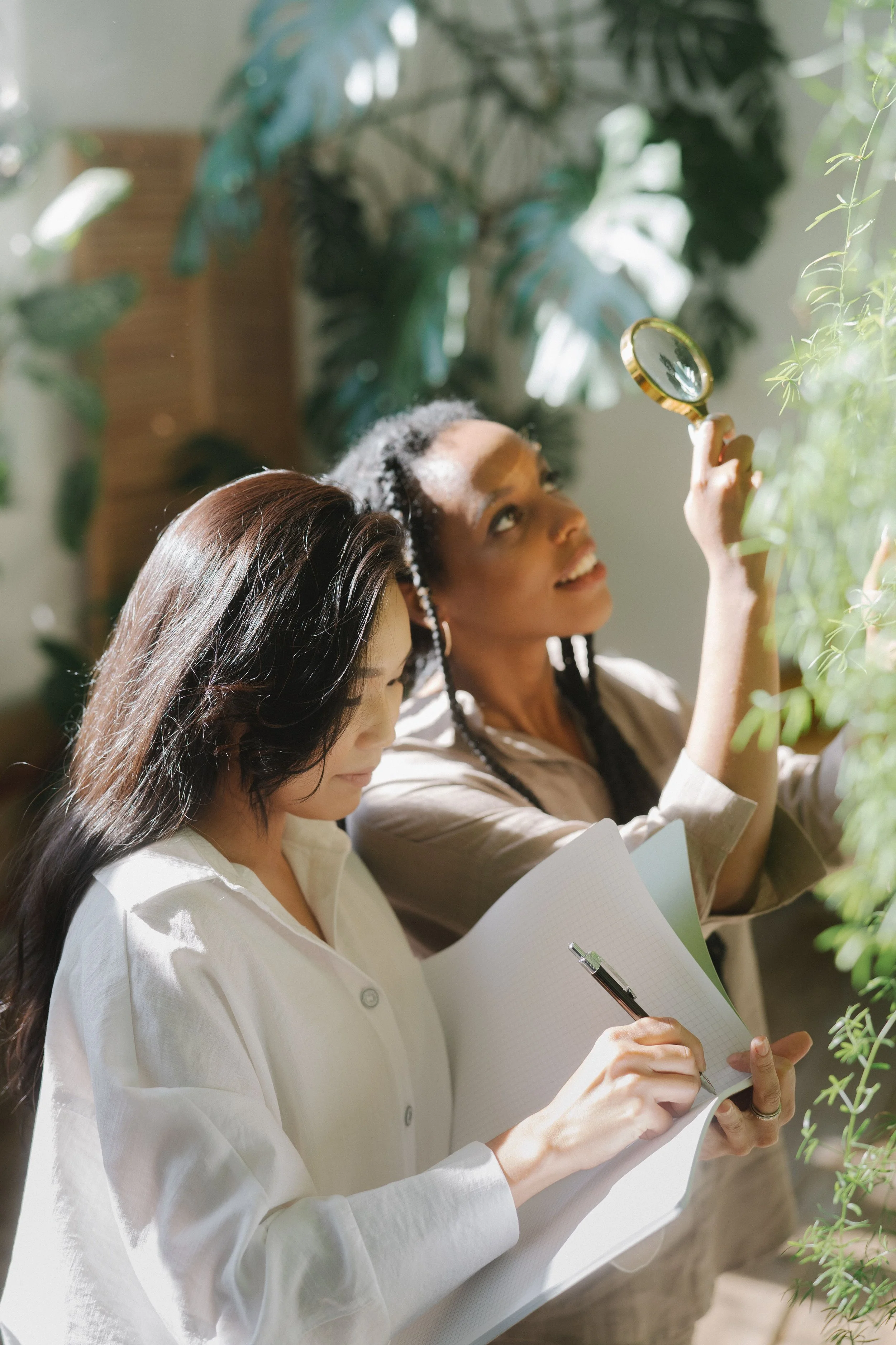 Two young women immersed in curiosity and connection, one jotting down discoveries in a journal while the other searches through a magnifying glass during a playful social adventure.