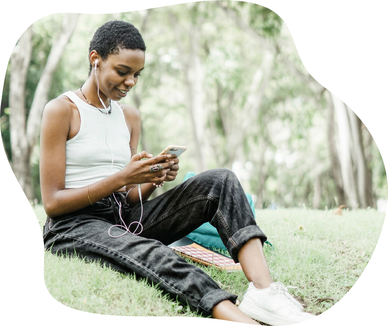 Young woman with short curly hair sitting on grass in a park, smiling while looking at her smartphone, with earphones in, wearing a white tank top and black jeans and doing a fun mindfulness activity for self-discovery.