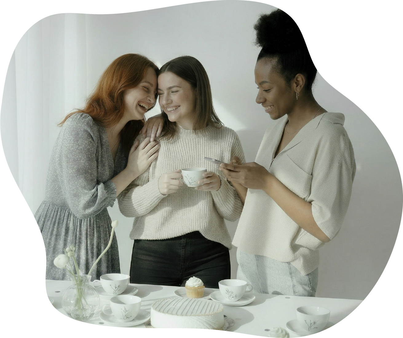Three women smiling and laughing around a table with tea cups, a cupcake, and a cake, enjoying a playful sensory mindfulness activity for girls night.