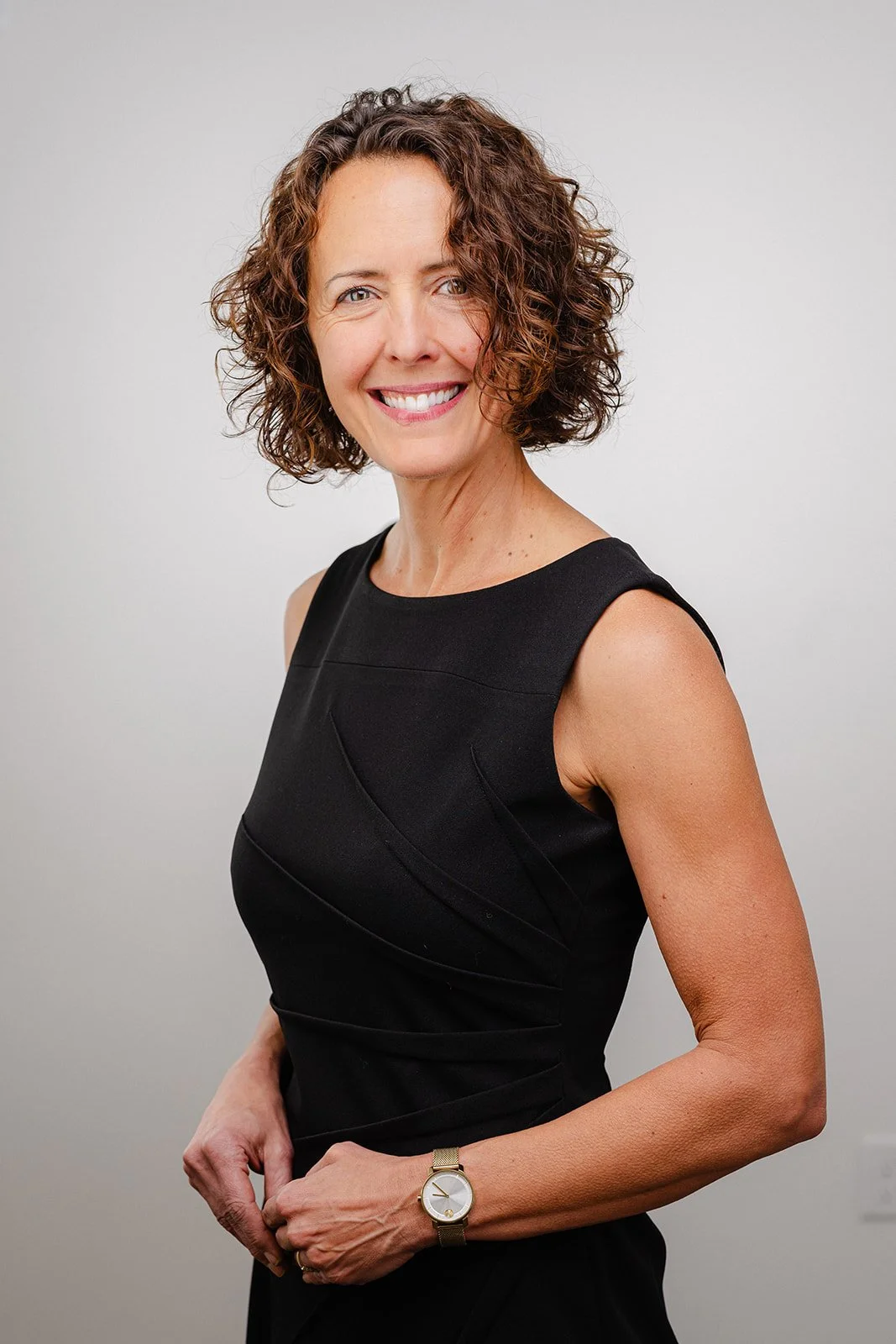 A smiling woman with curly brown hair wearing a sleeveless black dress and a gold watch against a plain light gray background.