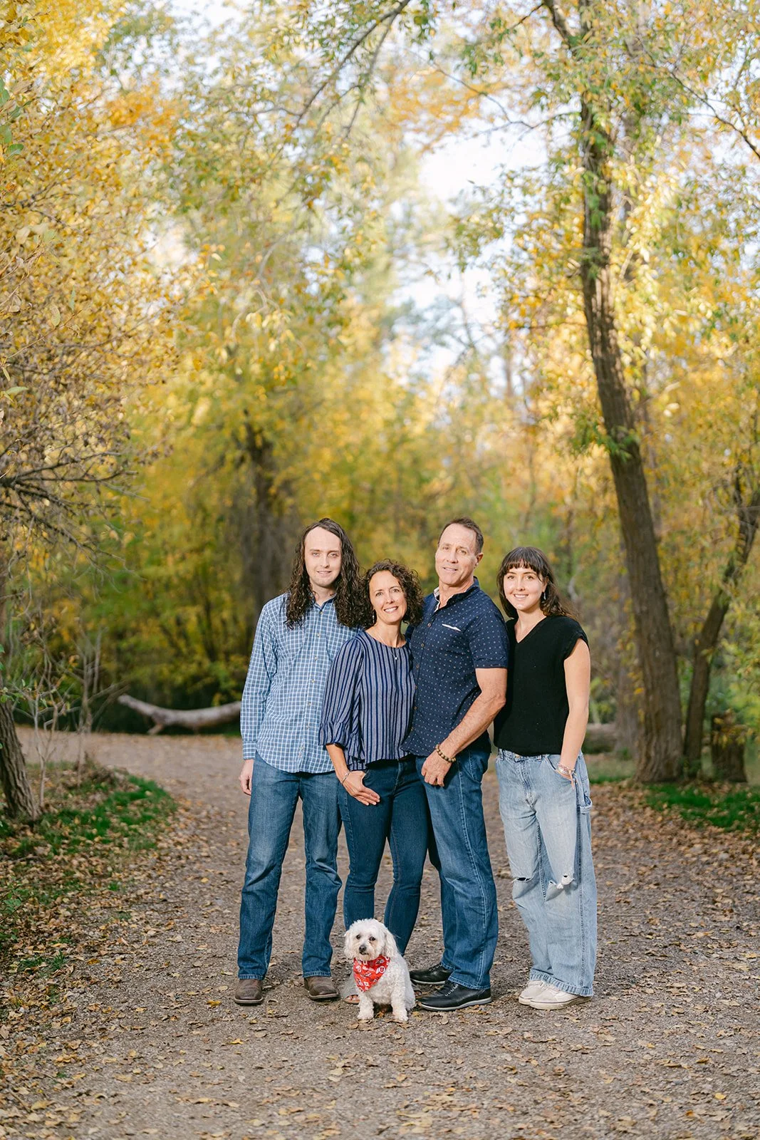 A family of four and a dog standing on a wooded path during fall with yellow and green leaves.