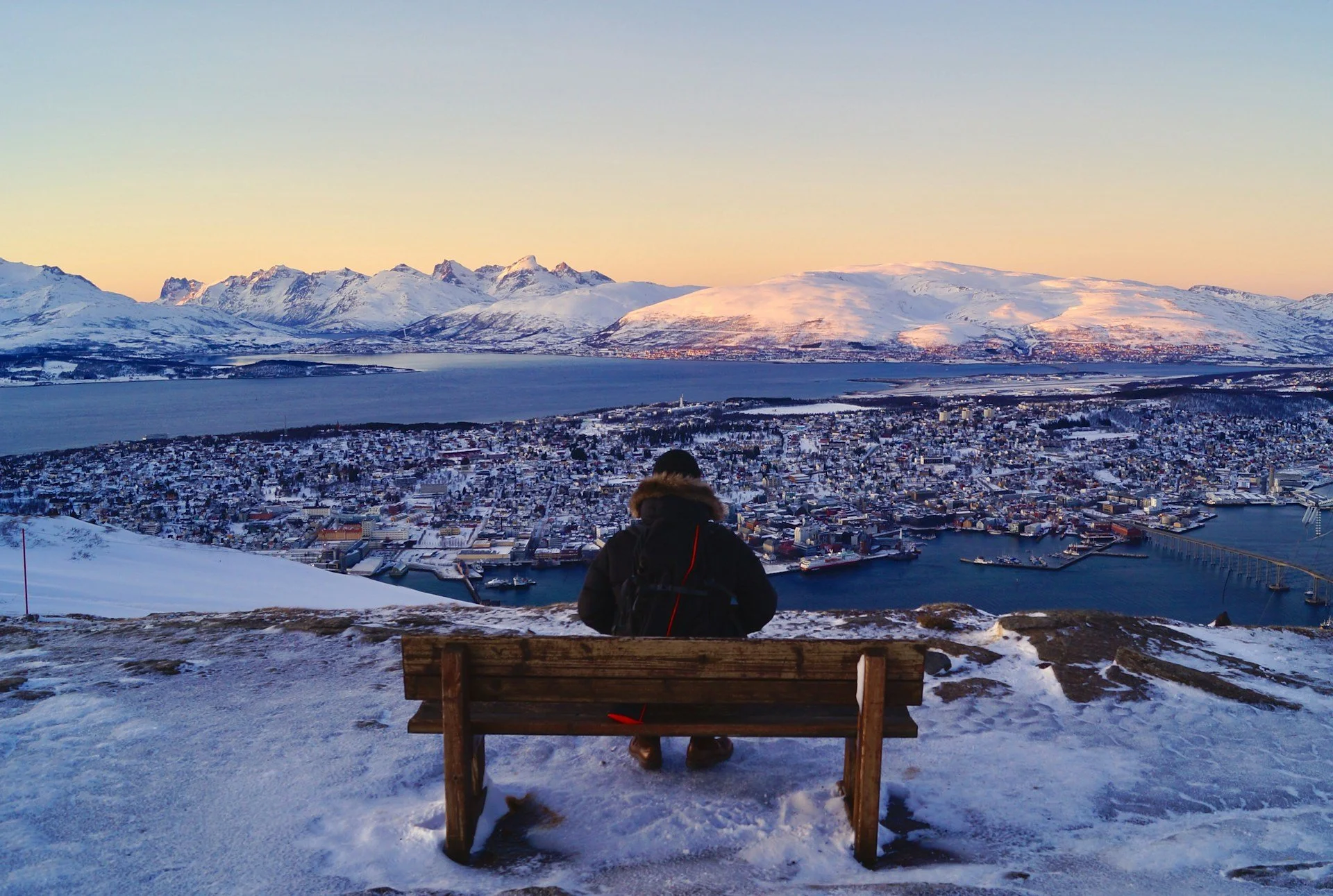 Person sitting on a bench overlooking a snowy mountain city and fjord at sunset.