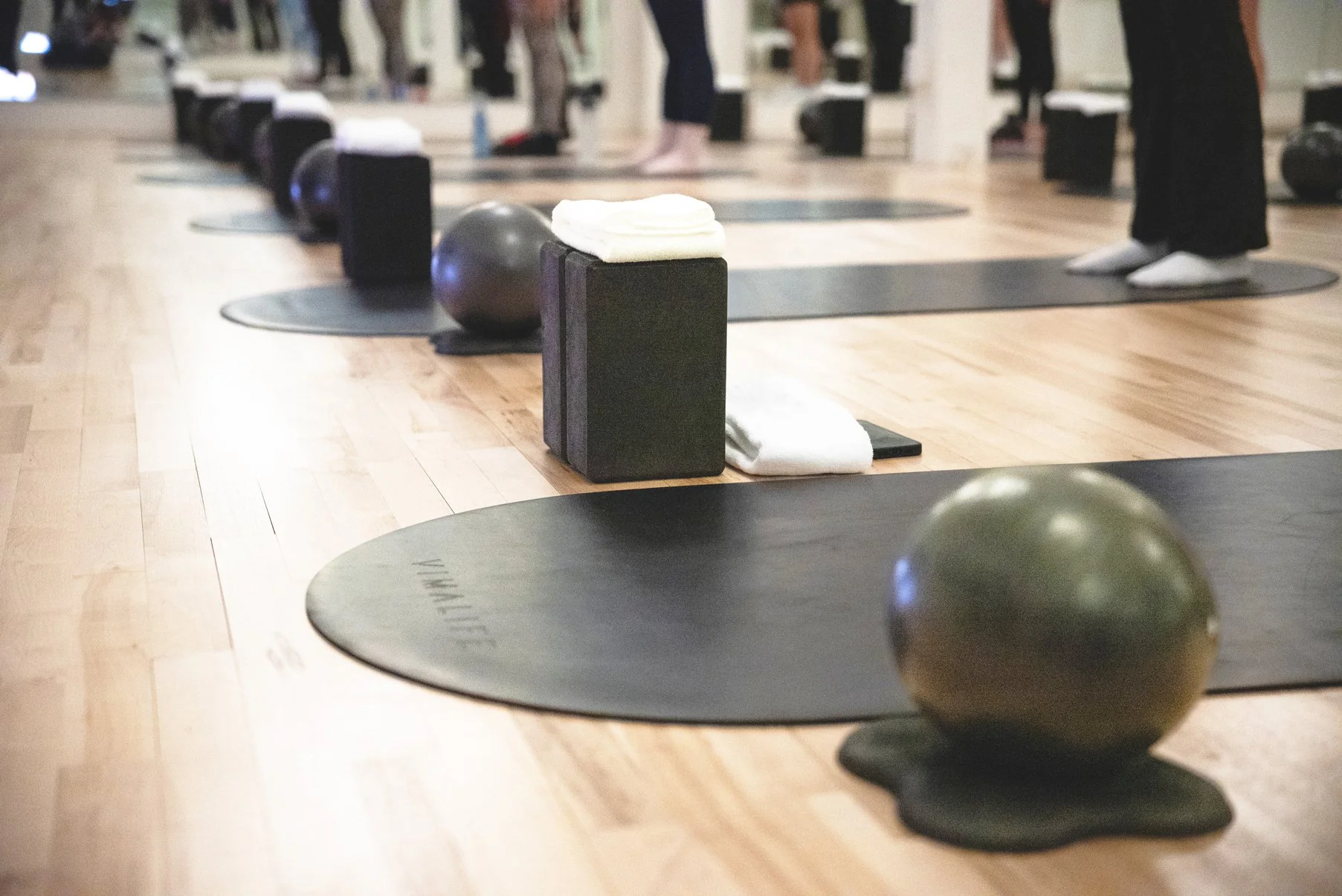 Yoga studio with black mats, black and gray exercise blocks, black exercise balls, and people standing in yoga poses, with towels and water bottles nearby.