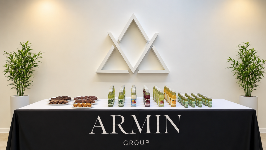 Table with snacks and drinks, branded with Armin Group logo, in a modern office space with two potted plants.