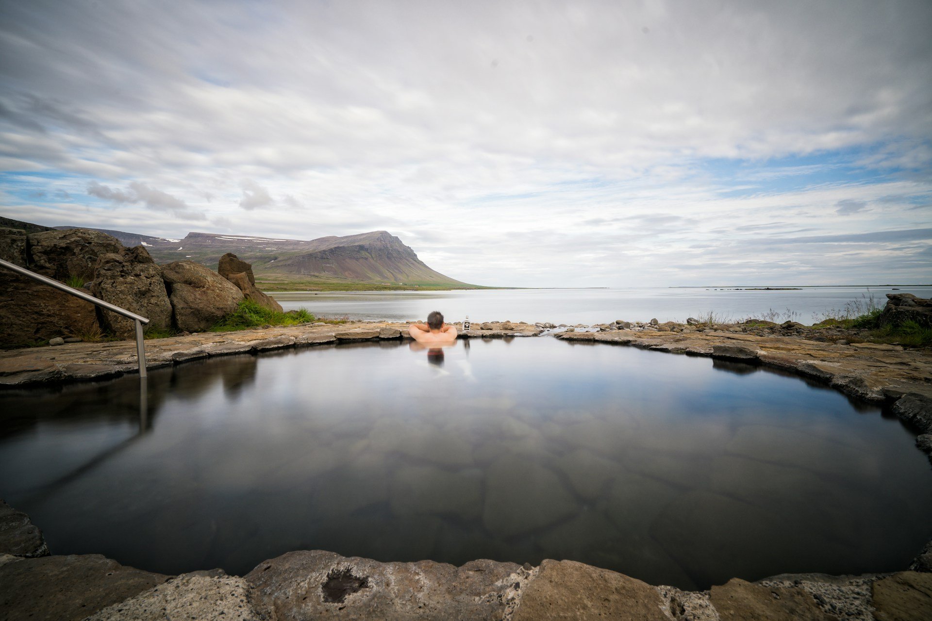 Person relaxing in an outdoor geothermal hot spring overlooking a mountain landscape.