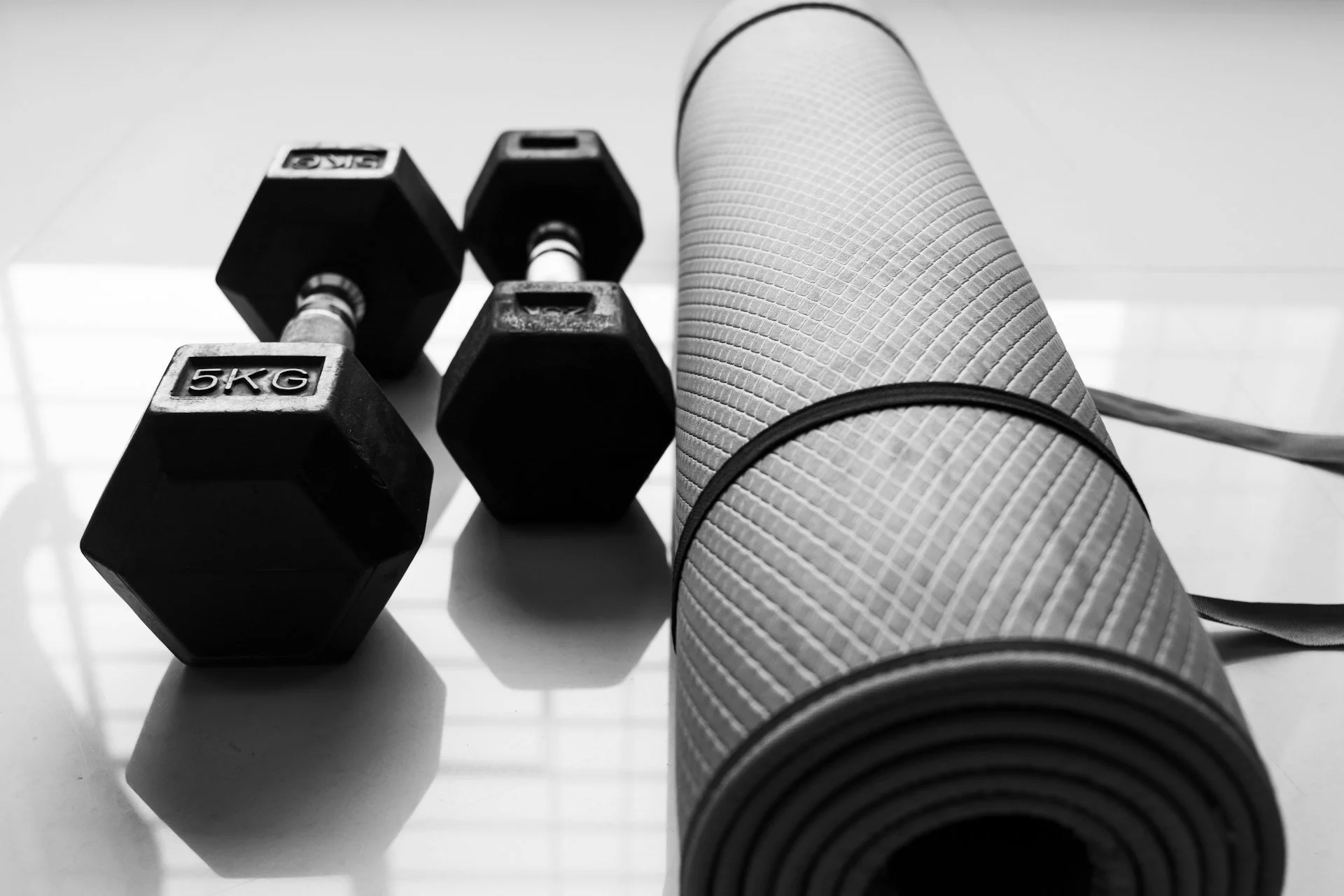 Black-and-white photo of dumbbells beside a rolled exercise mat for strength and mobility training.
