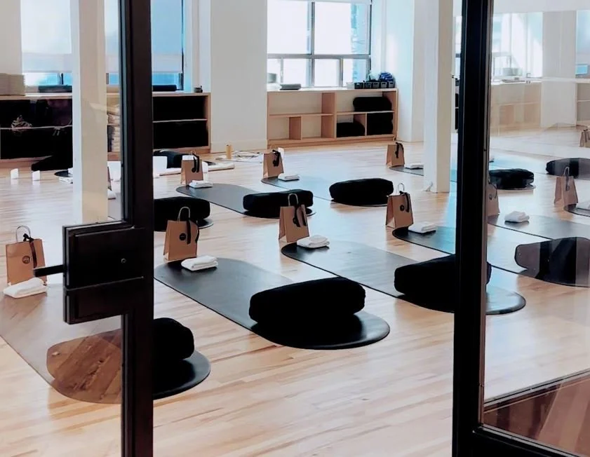 Yoga studio with black mats, bolsters, and folded towels on either side, set up for a class, with a bright, open space and large windows.