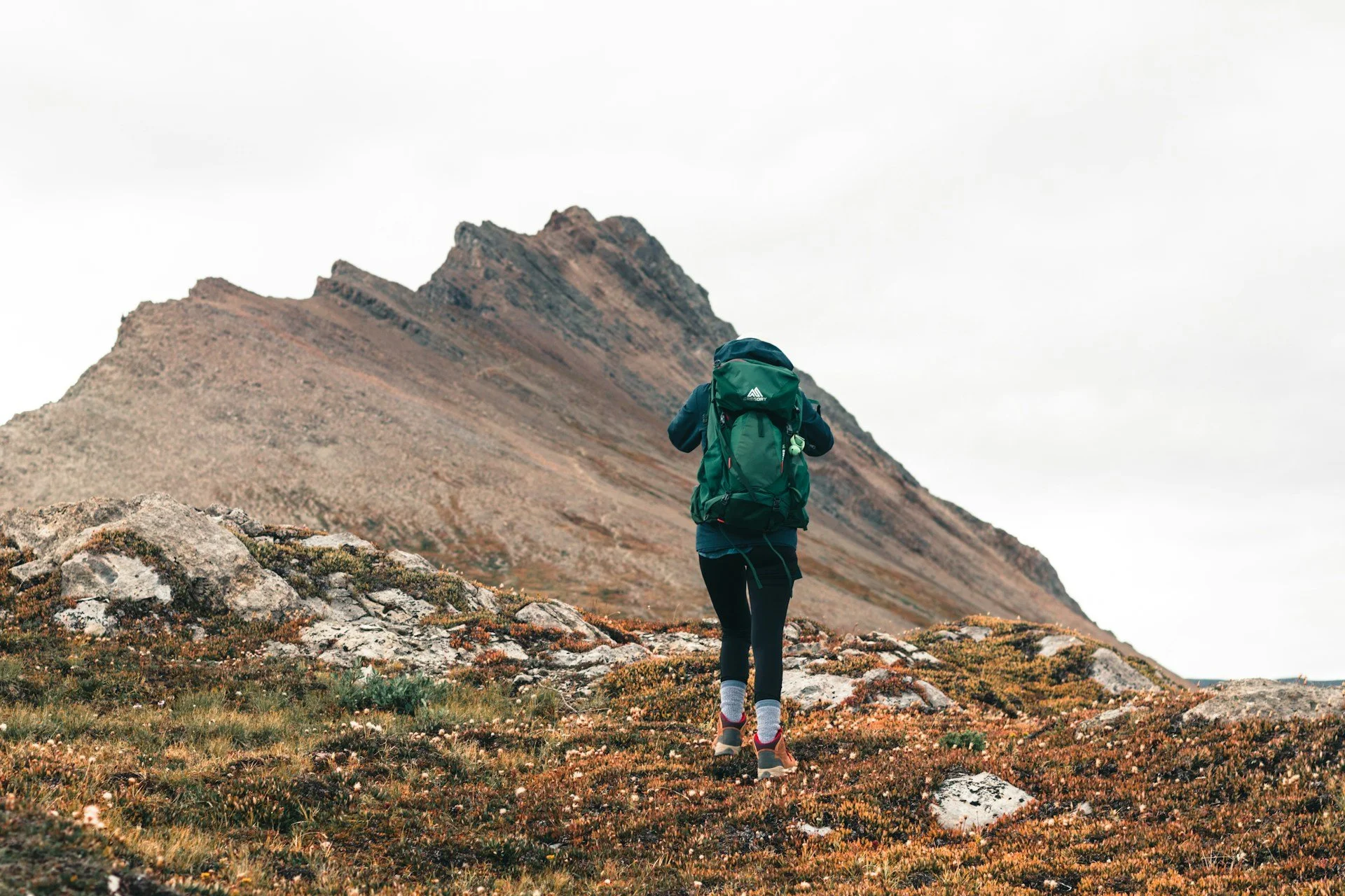 Hiker carrying a weighted backpack toward a rocky mountain peak.