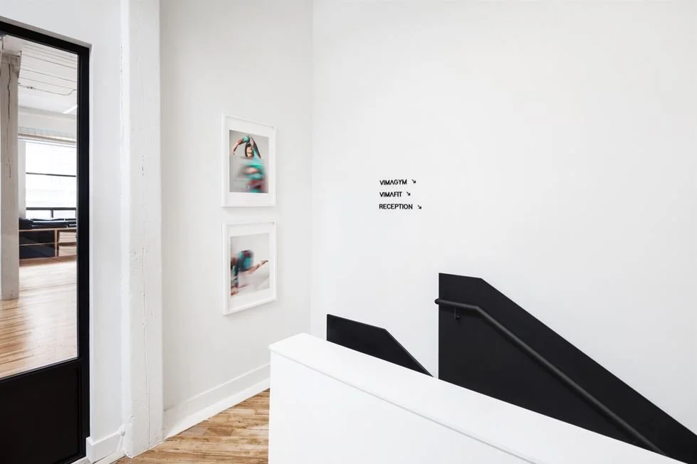 Interior staircase with white walls, black handrail, two blurred art photos on the wall, and door leading to a room with wooden floors.