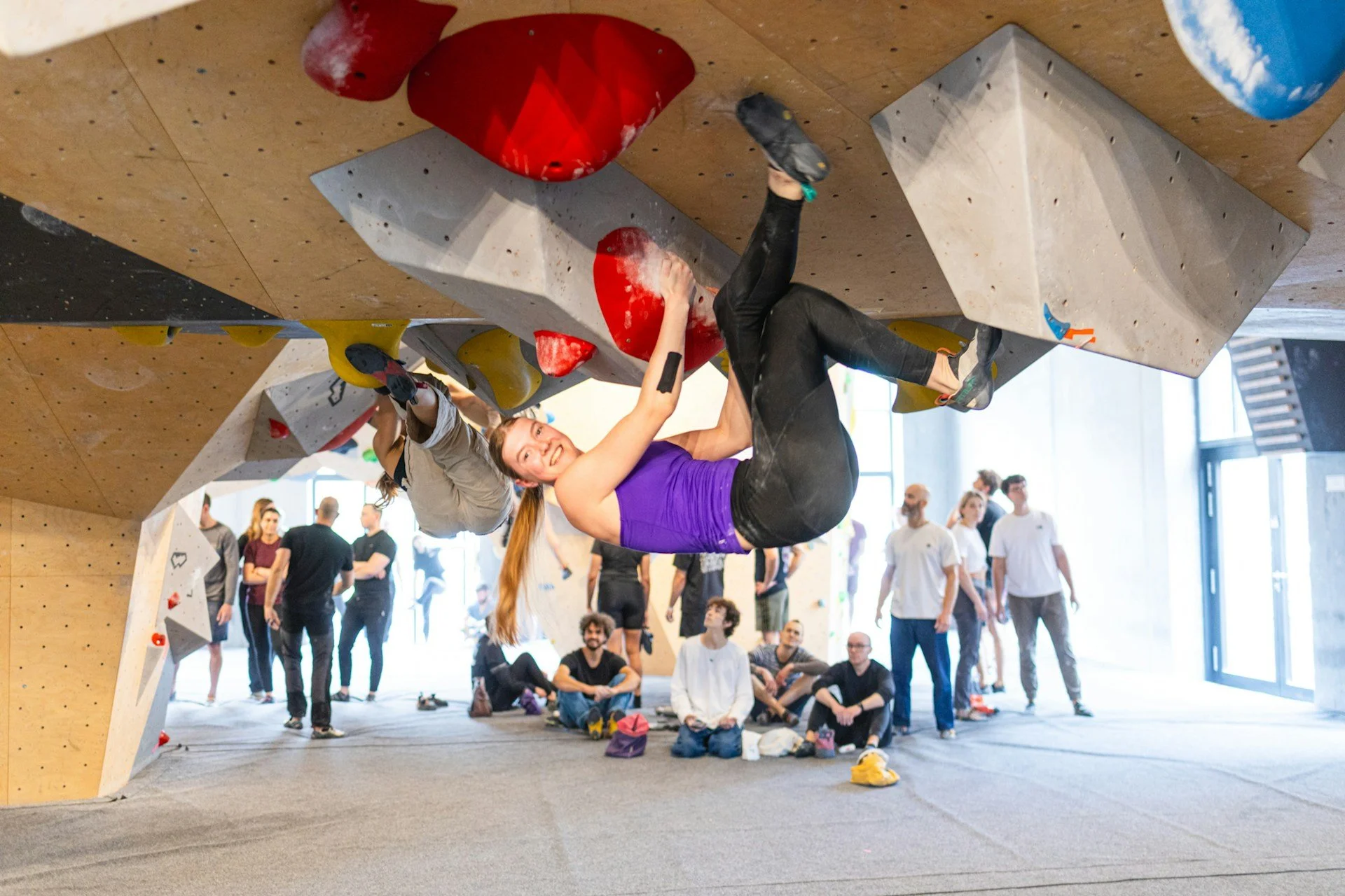 Indoor bouldering climber on an overhang wall with spectators watching.