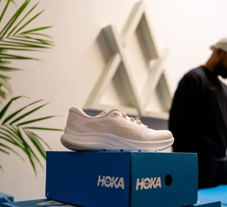 A white athletic shoe displayed on a blue box with the HOKA logo in a retail store, with a blurred person in black apparel and a plant in the background.