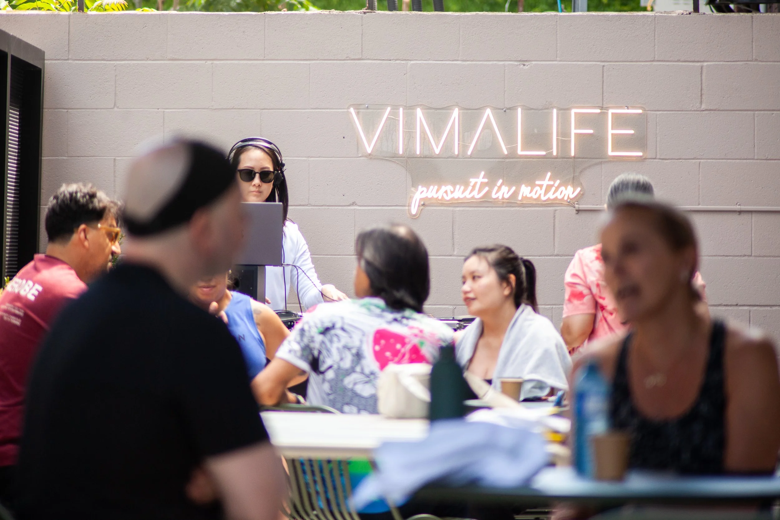 VIMALIFE members relaxing at a wellness event with a DJ and neon pursuit in motion sign.