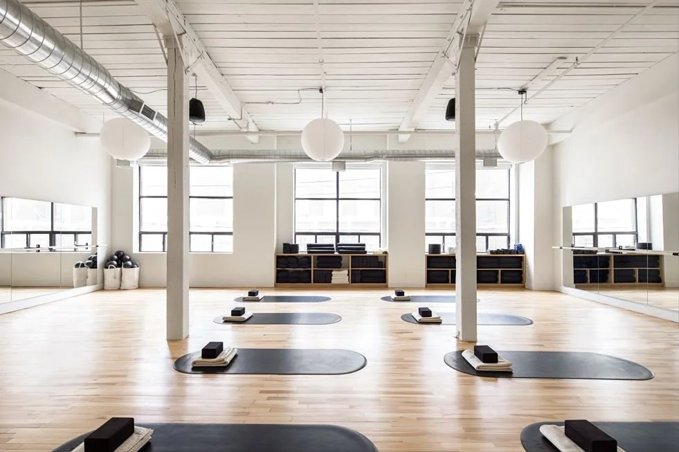 Empty yoga studio with mats and blocks set up for practice, mirrored wall, large windows, and minimal decor.
