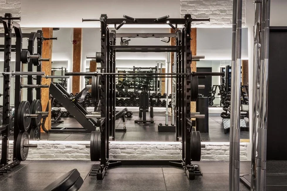 A gym with weightlifting equipment, including barbells, weight plates, and a squat rack, with mirrors and brick walls.