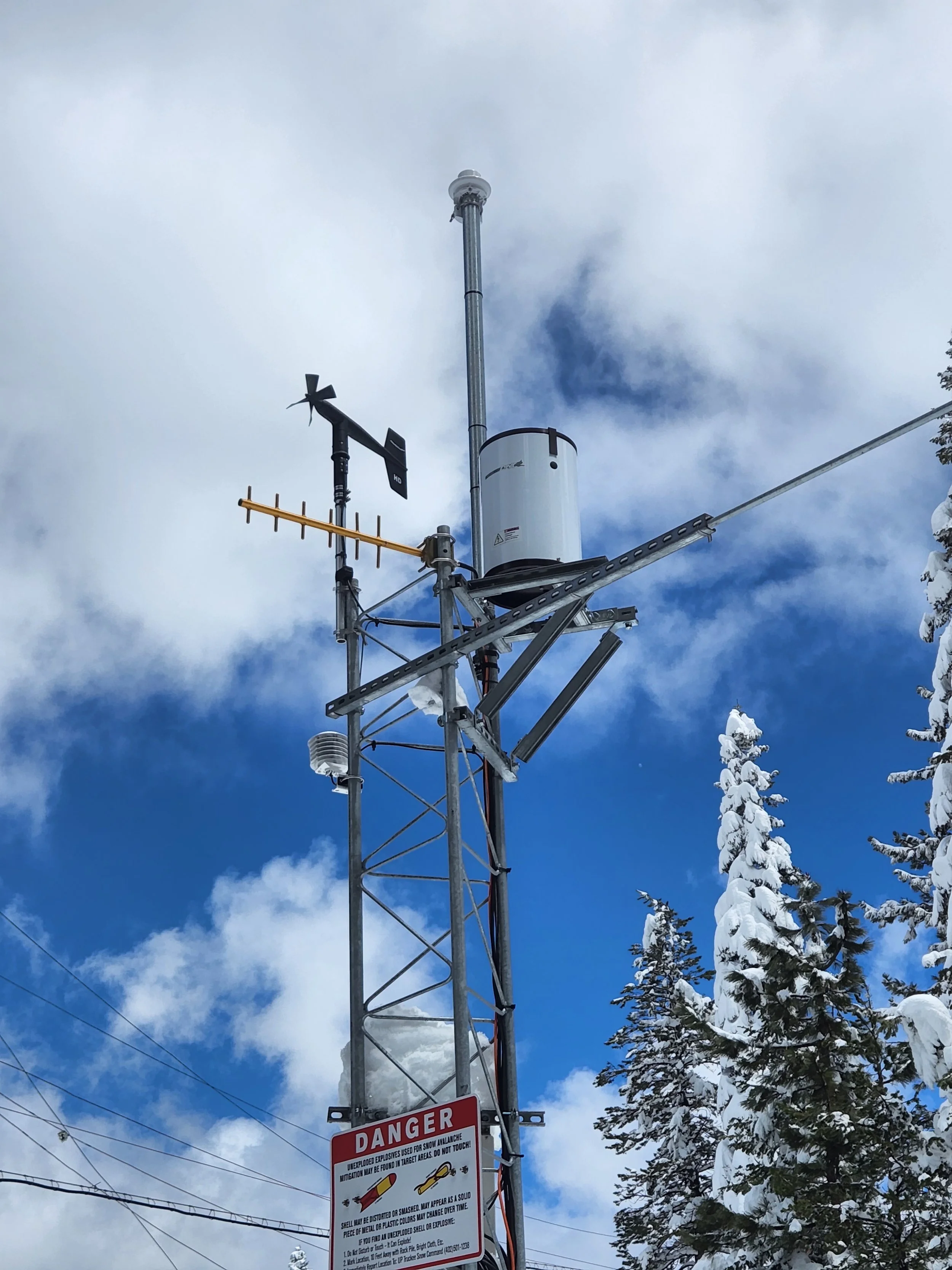 A weather station with antennas and sensors on a metal tower against a winter sky with snow-covered trees.