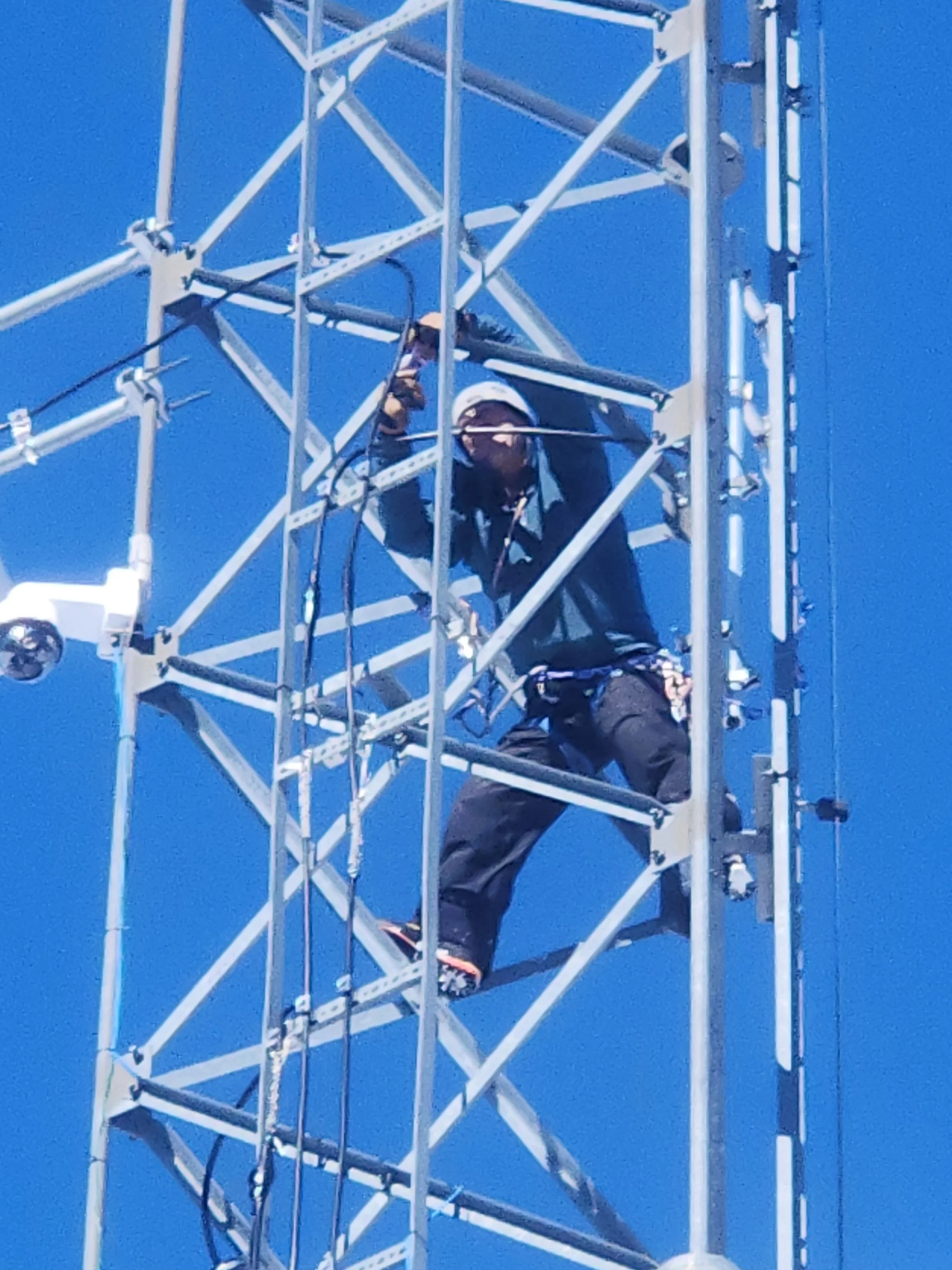 A worker inspecting or repairing a radio tower while perched on its metal framework against a clear blue sky.