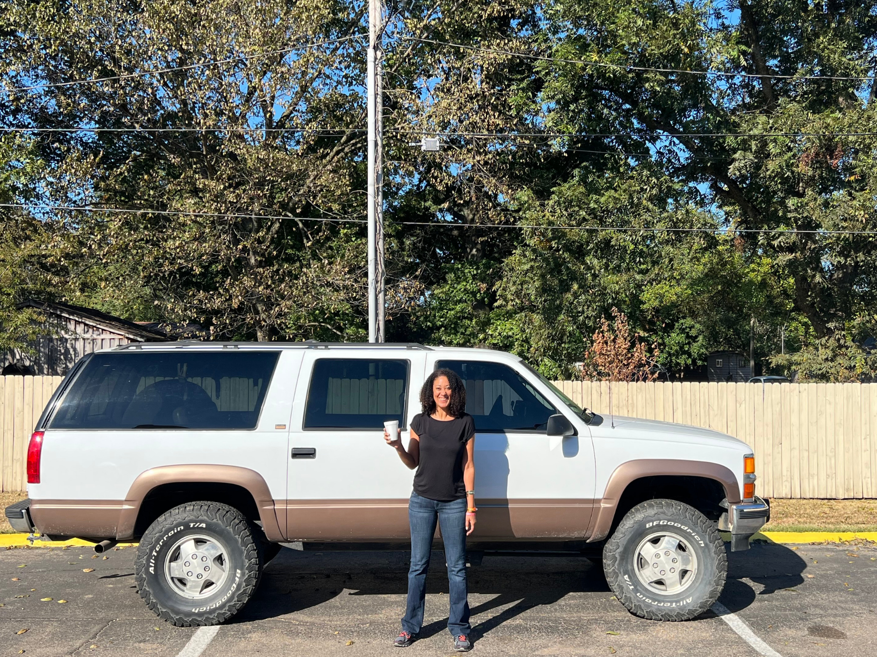 A woman standing in front of a white and brown SUV, holding a drink and smiling outdoors on a sunny day with green trees and a wooden fence in the background.