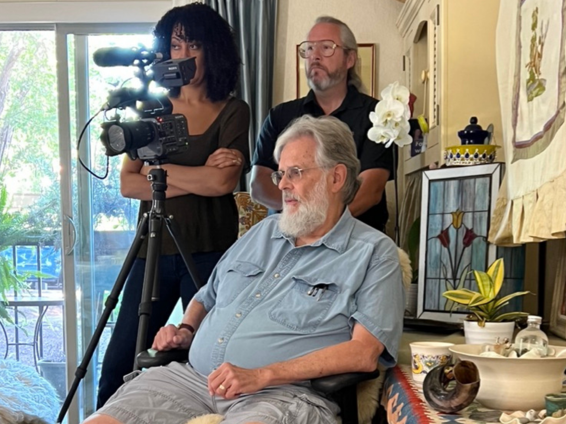 An elderly man with glasses and a beard sits in a wheelchair during a filming or photography session. Behind him, two people, a woman with curly dark hair and a man with gray hair and glasses, observe the scene. The woman is operating a video camera 