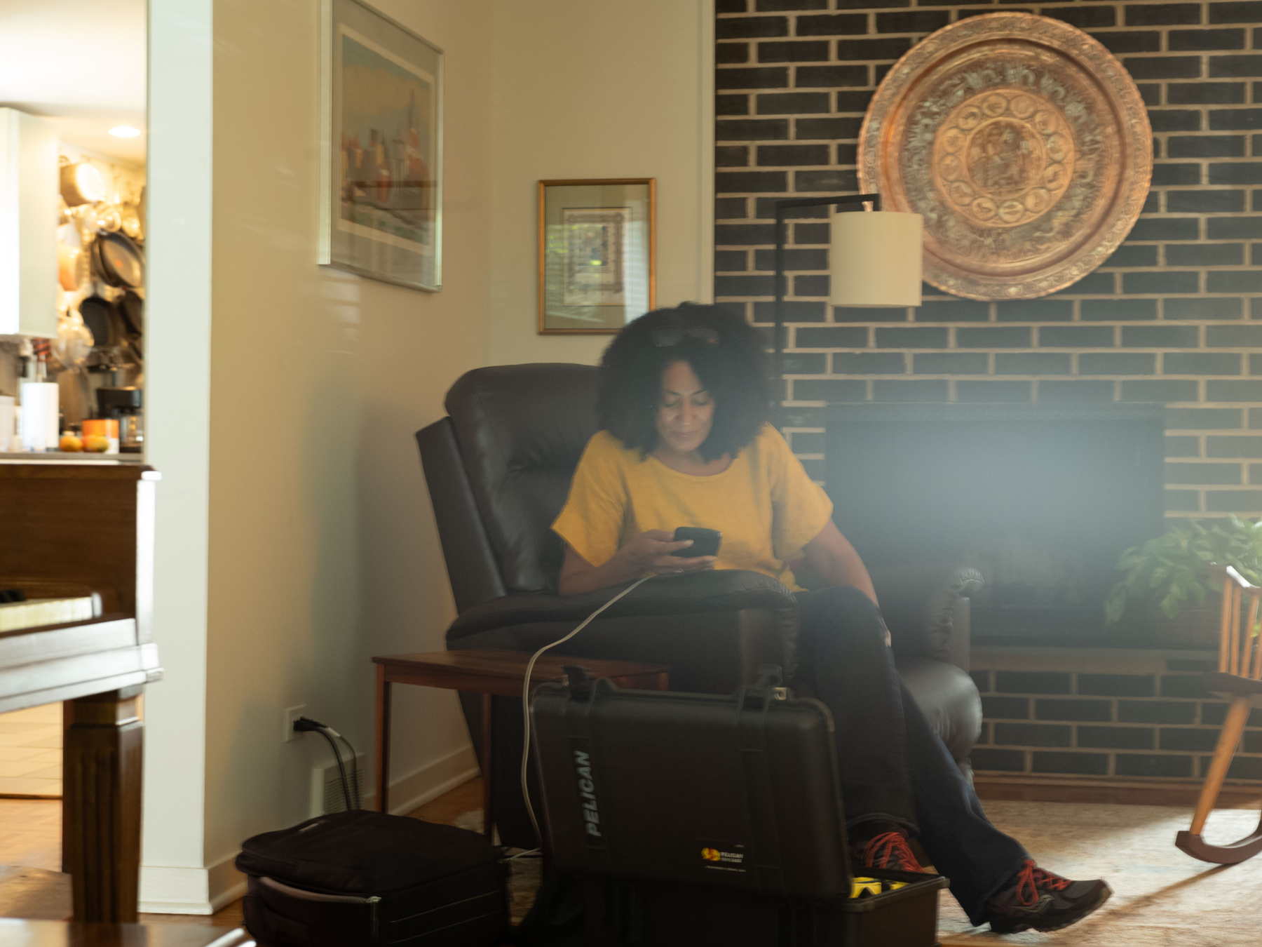 Woman with curly black hair wearing a yellow t-shirt sitting in a recliner, looking at her phone, in a living room with brick wall, framed pictures, and a potted plant.