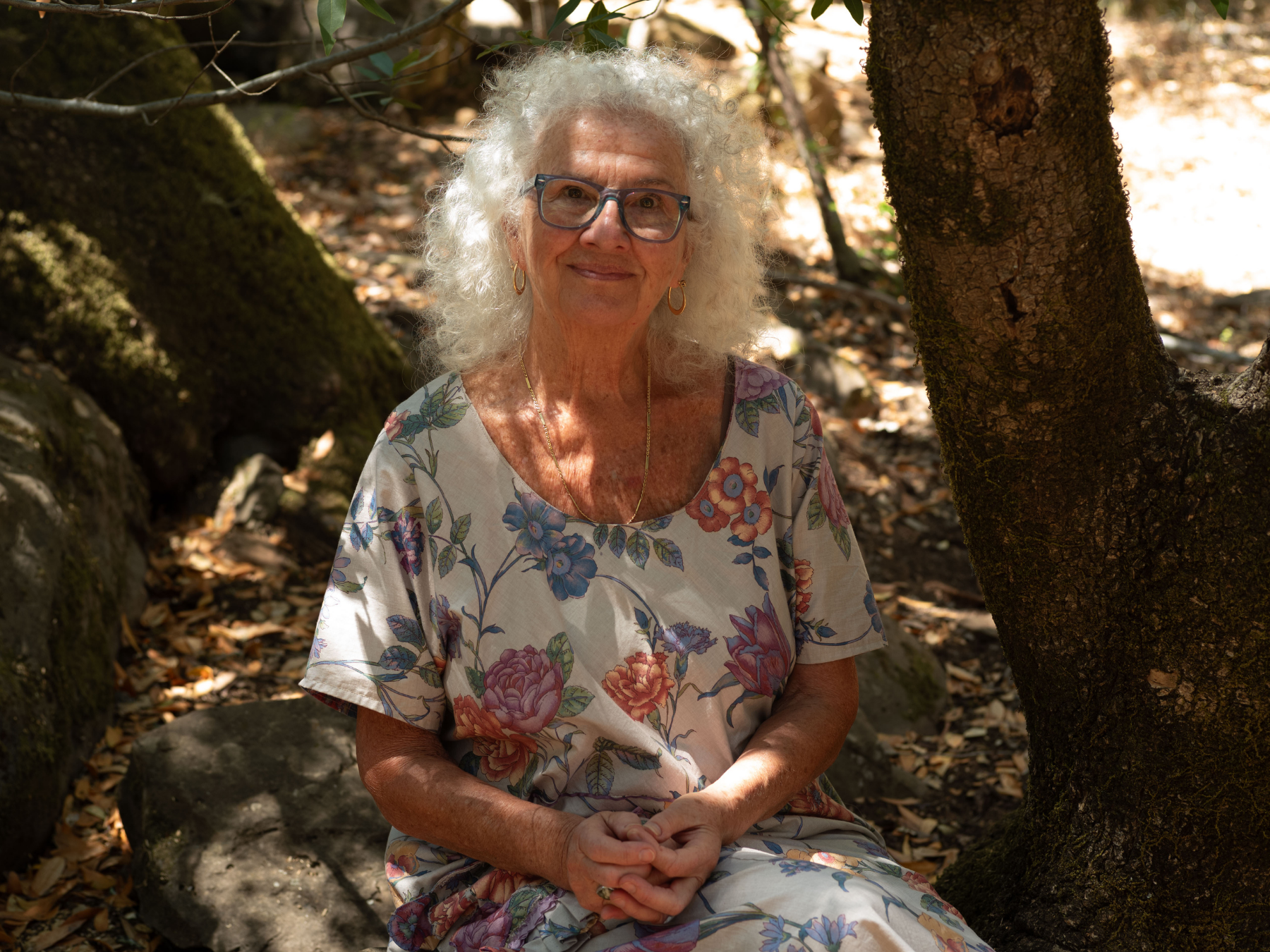 An elderly woman with curly white hair and glasses, wearing a floral dress, sitting outdoors in a wooded area with sunlight filtering through the trees.