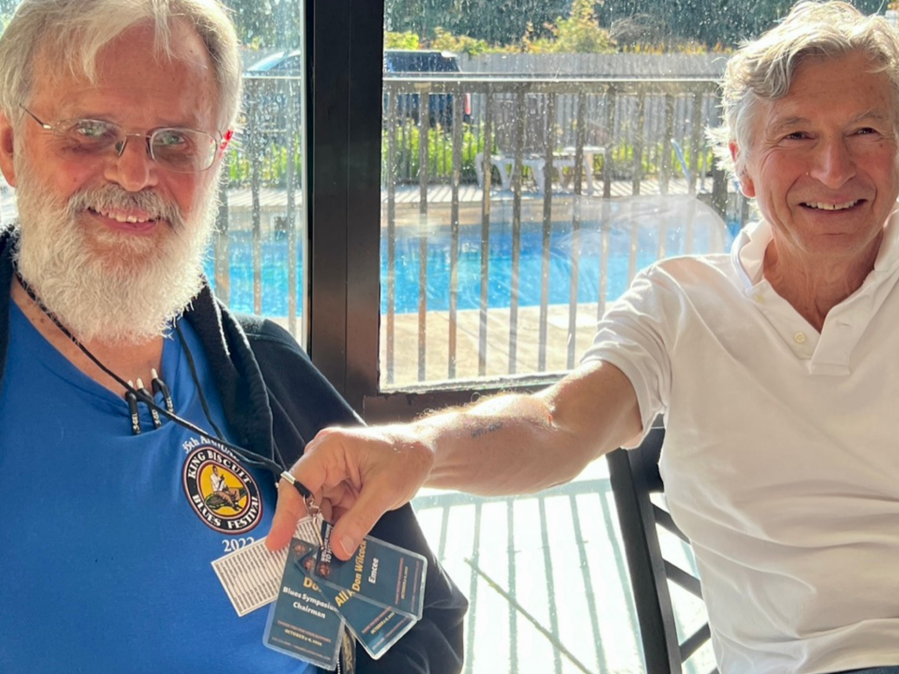 Two elderly men smiling, one with a white beard and glasses wearing a blue shirt with a badge, and the other with gray hair in a white polo shirt, shaking hands inside a room with glass doors showing a pool outside.