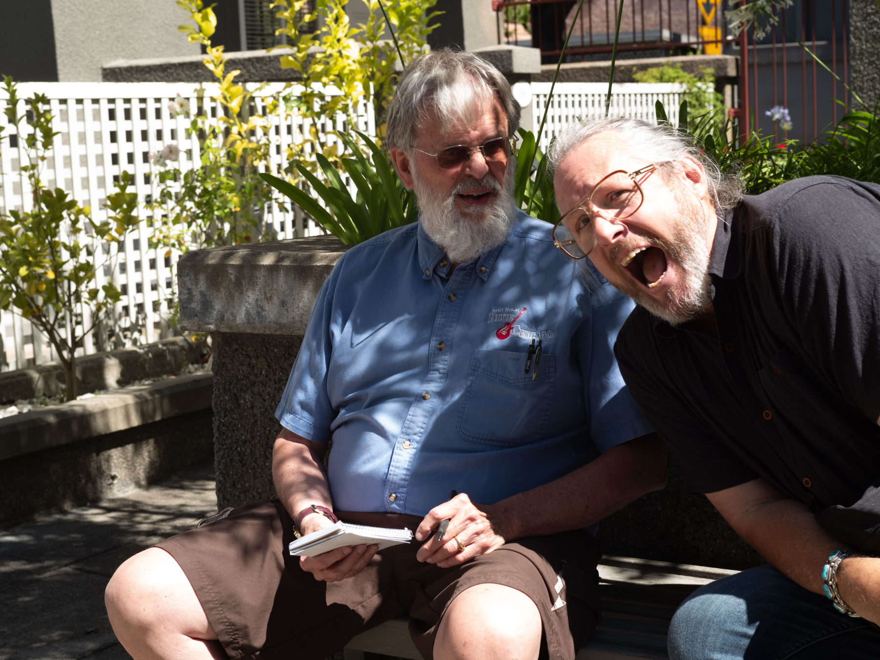 Two older men are sitting on a park bench outdoors, engaged in a lively conversation and laughing. One man is wearing a blue shirt with tools in the pocket, and the other is wearing a dark shirt and glasses. There are plants and a white fence in the 