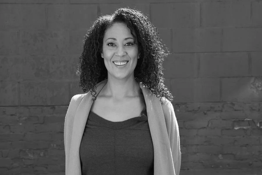 Smiling woman with curly hair standing in front of a textured brick wall.