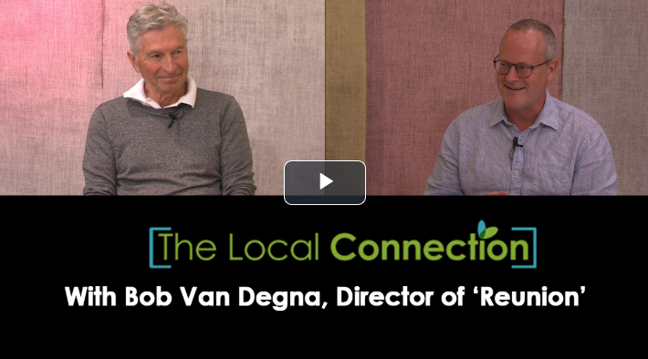 Two men sitting on stage during an interview or panel discussion. The background is pink and beige. The text overlay reads "The Local Connection" with Bob Van Degna, director of 'Reunion'.