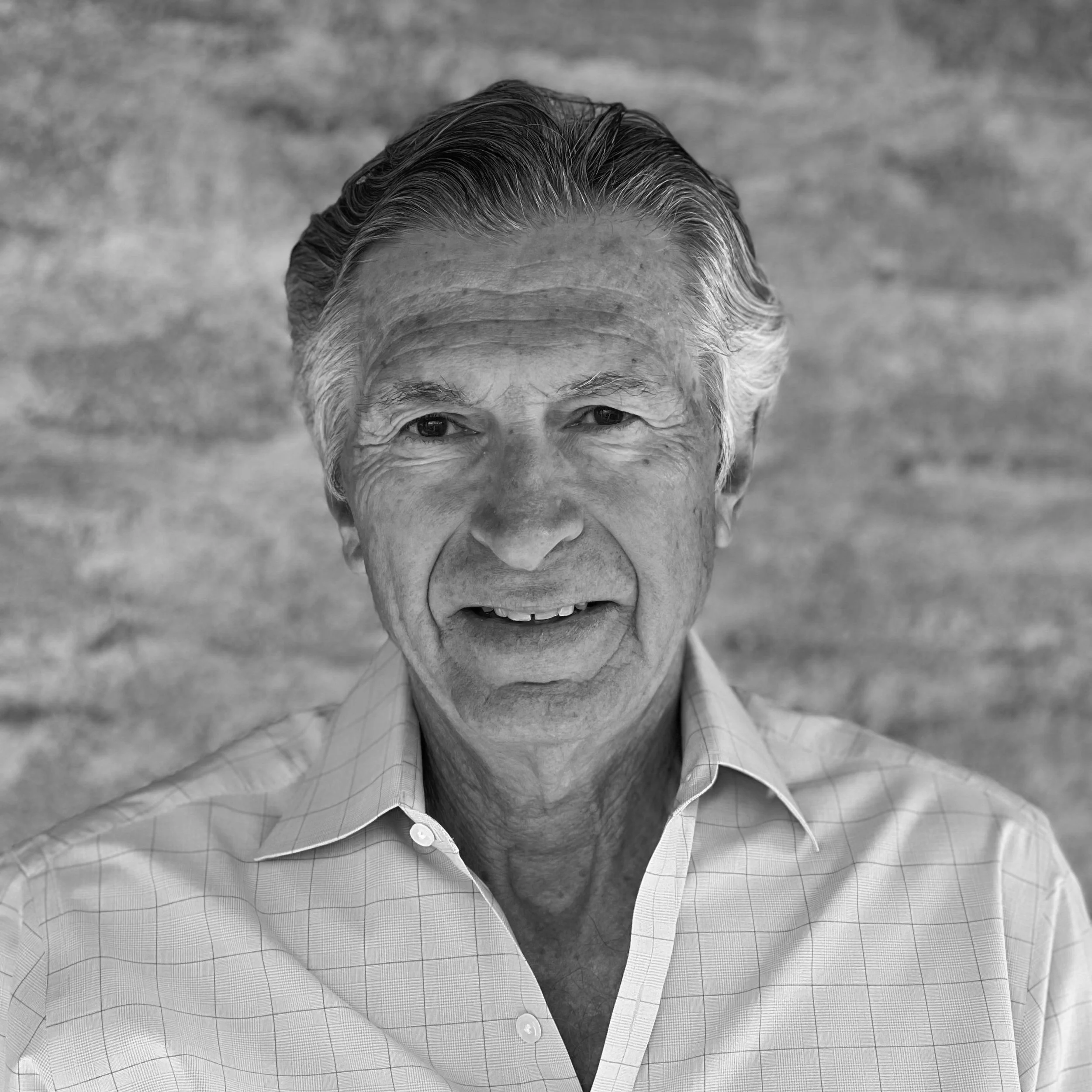 Black and white close-up photo of an elderly man with gray hair, wearing a collared shirt, standing outdoors against a blurred textured background.