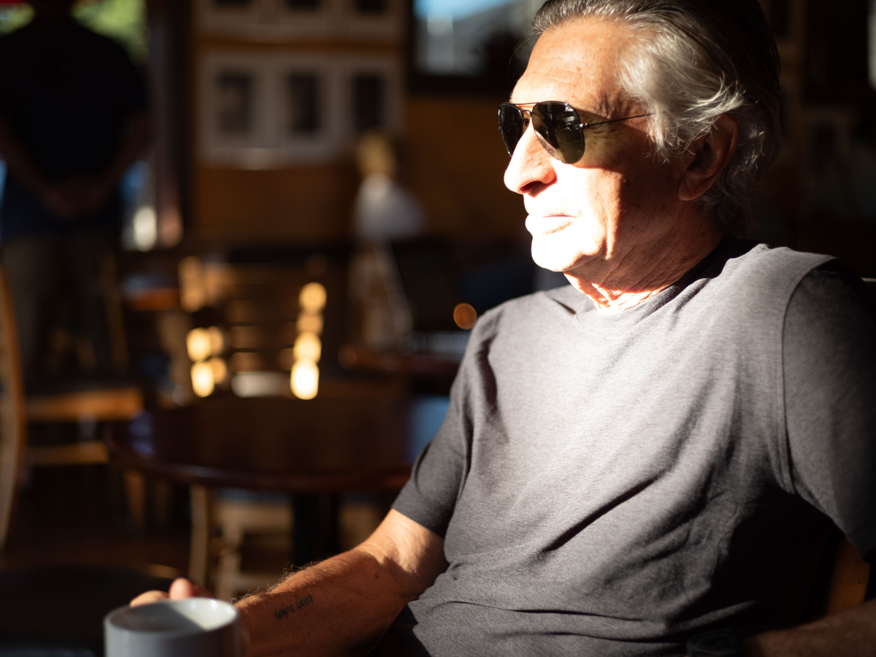 An elderly man with gray hair, wearing sunglasses and a gray t-shirt, sitting in a cafe with sunlight illuminating his face, holding a white mug, with blurred wooden chairs and tables in the background.